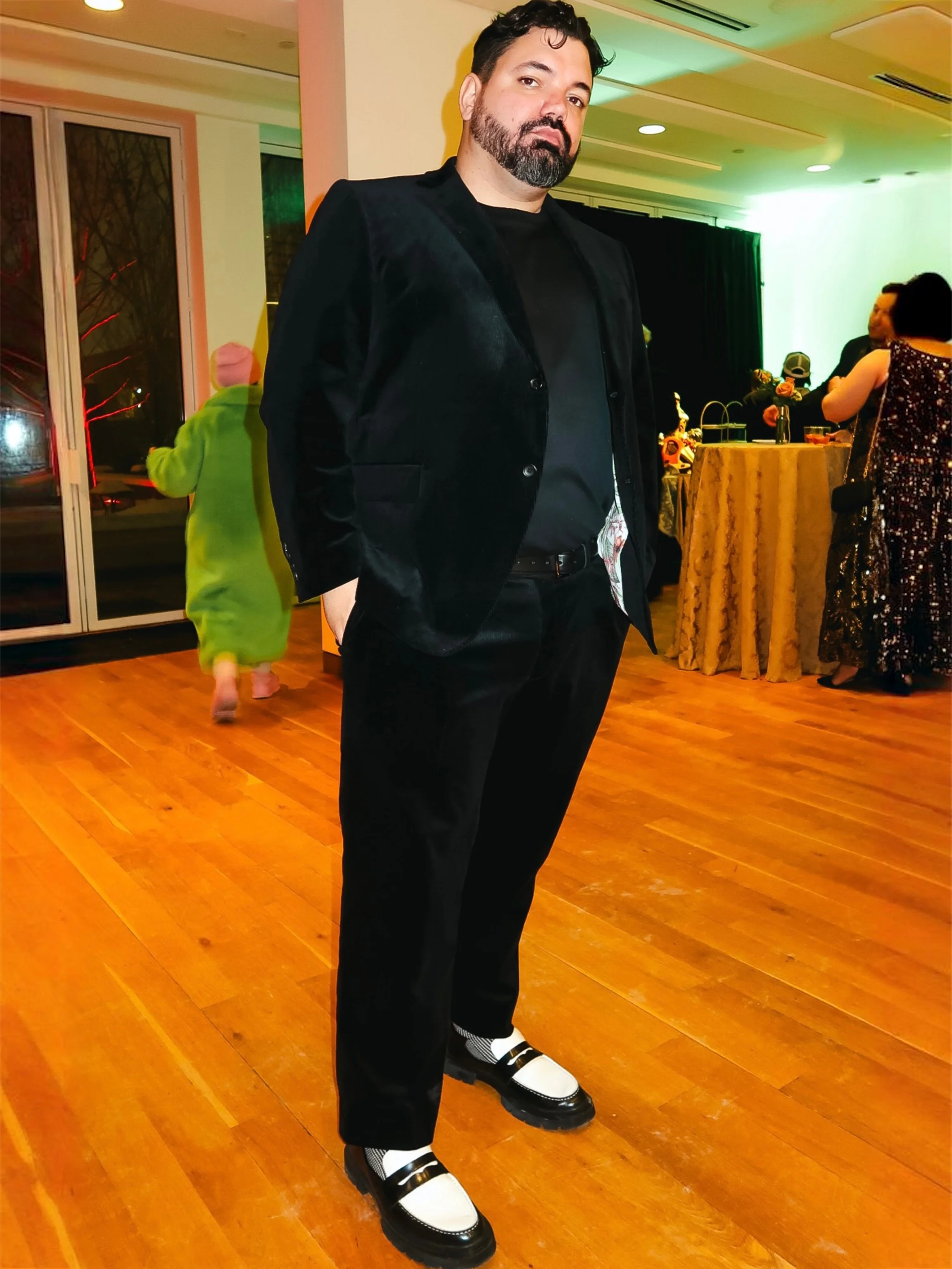 A man in formal black attire standing on a wooden floor at a social event, with people and decorated tables in the background.