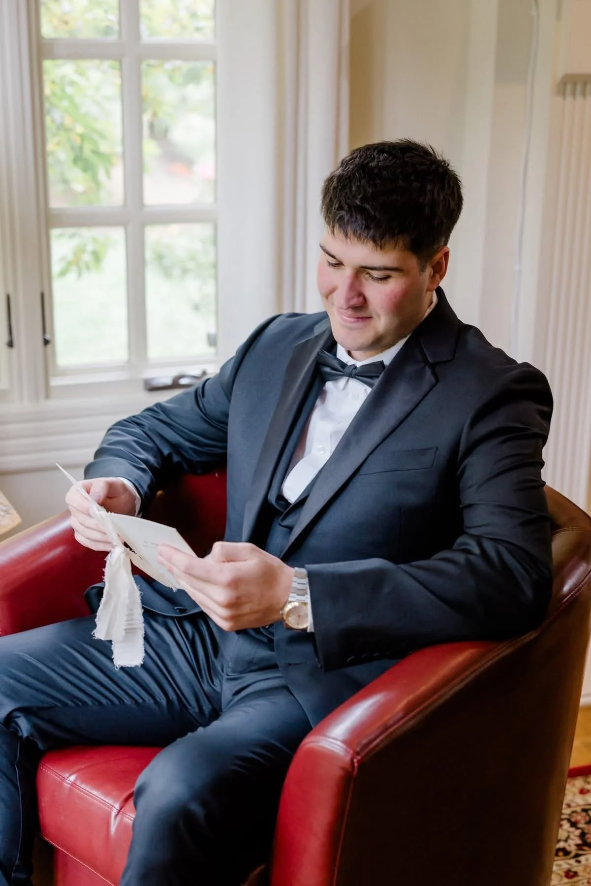 A young man dressed in a black tuxedo with a bow tie, sitting on a red leather chair, looking at and holding a card or letter. He is in a room with large windows and white curtains, bright natural light coming in.