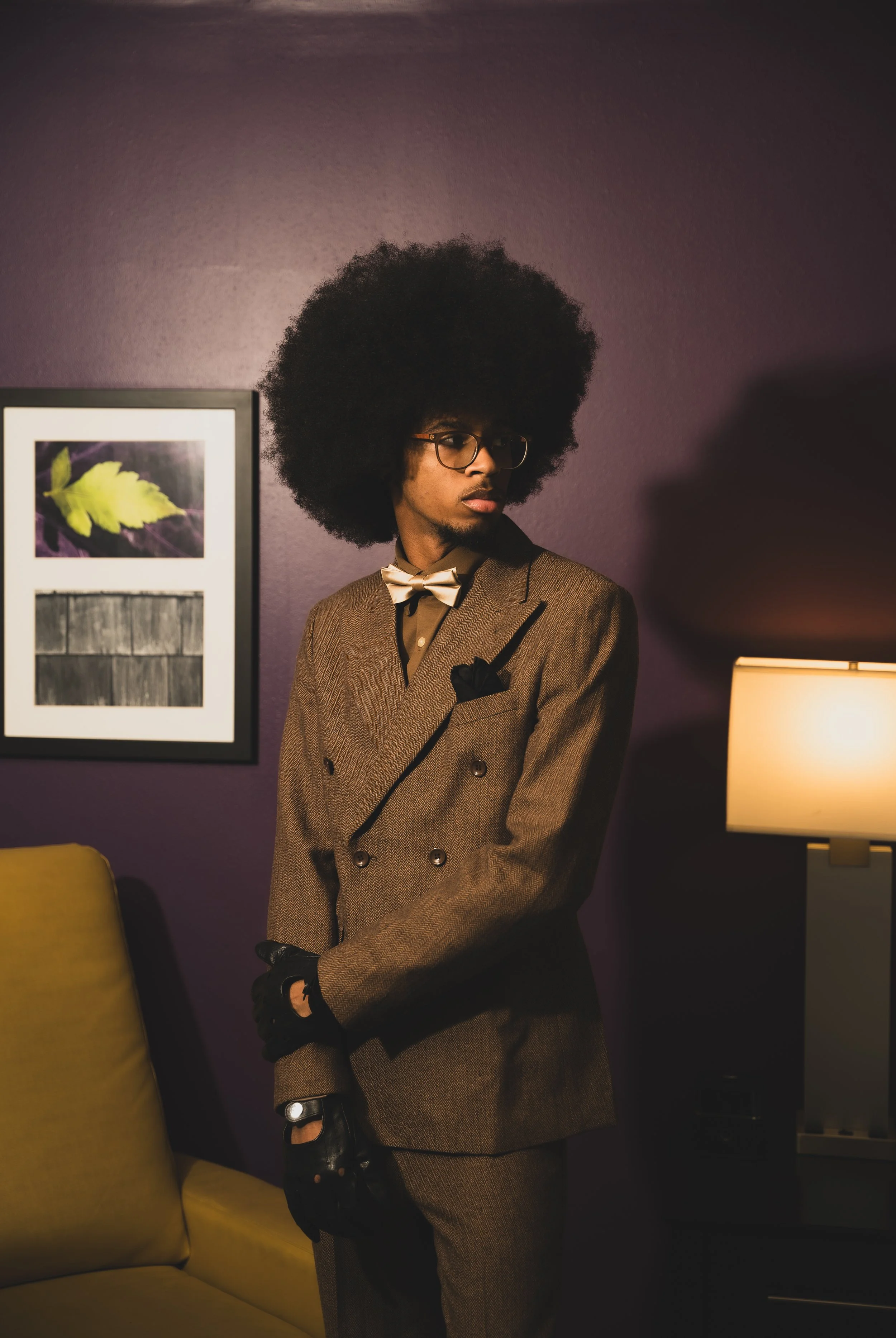 A man with a large afro hairstyle wearing glasses, a brown double-breasted suit, a bow tie, and black gloves, standing indoors against a purple wall with framed pictures and a lamp.