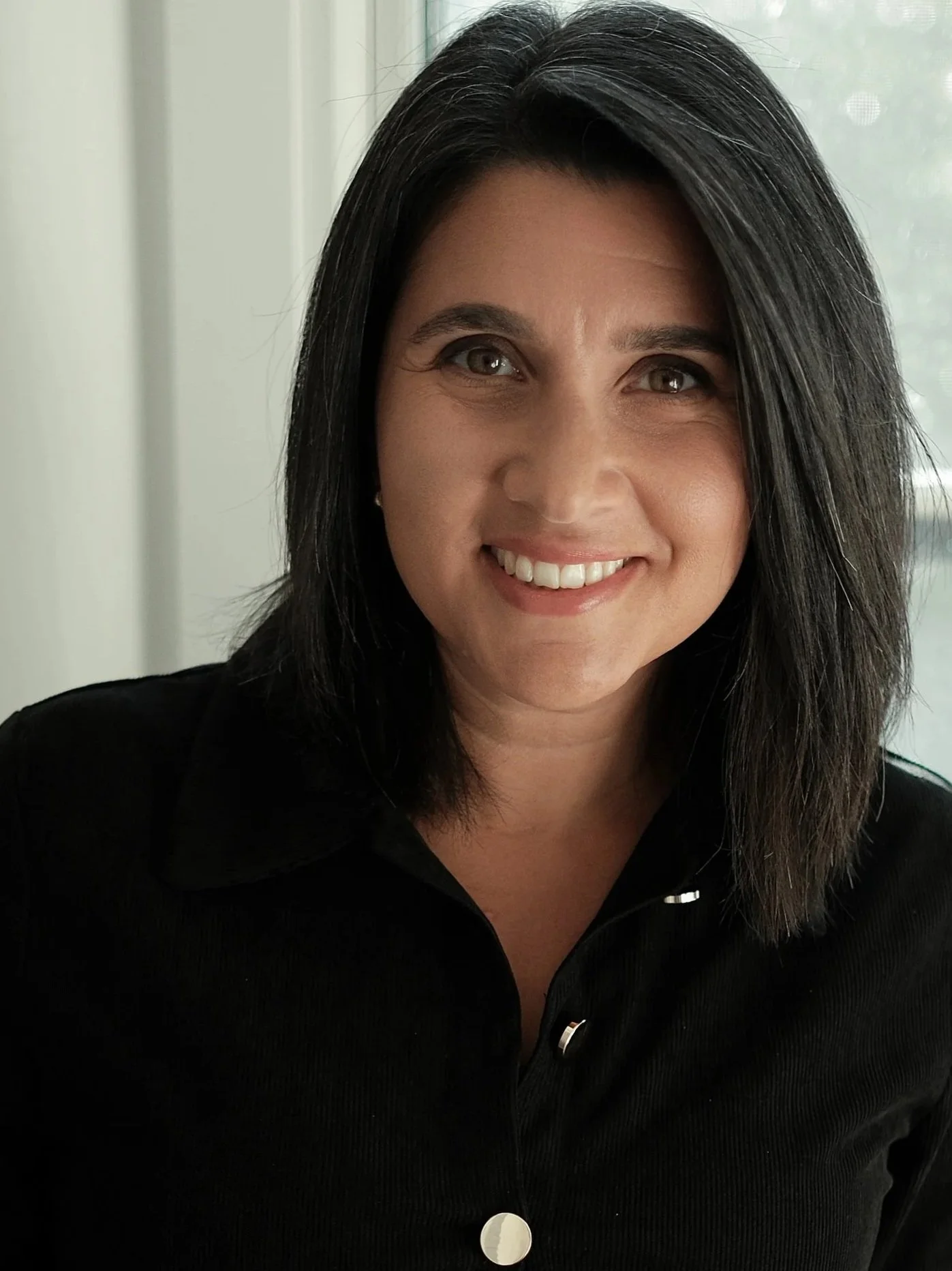 A woman with black hair, wearing a black collared shirt, smiling in front of a light background with a window.