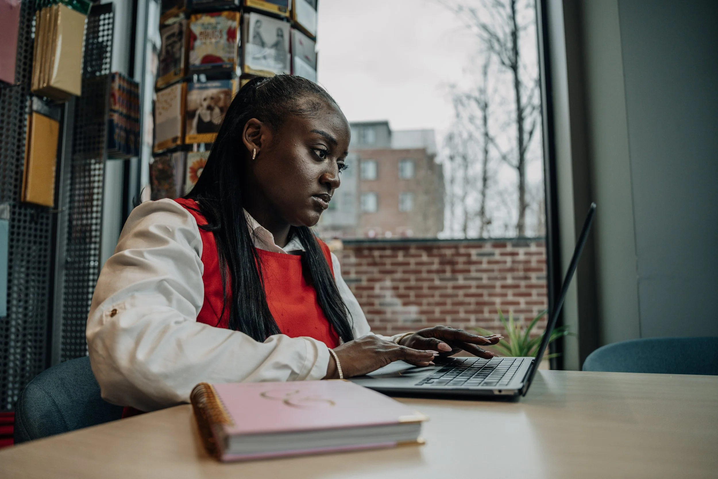 A woman wearing a red apron working on a laptop in a cozy indoor setting with a large window showing an outdoor scene with trees and a brick building.