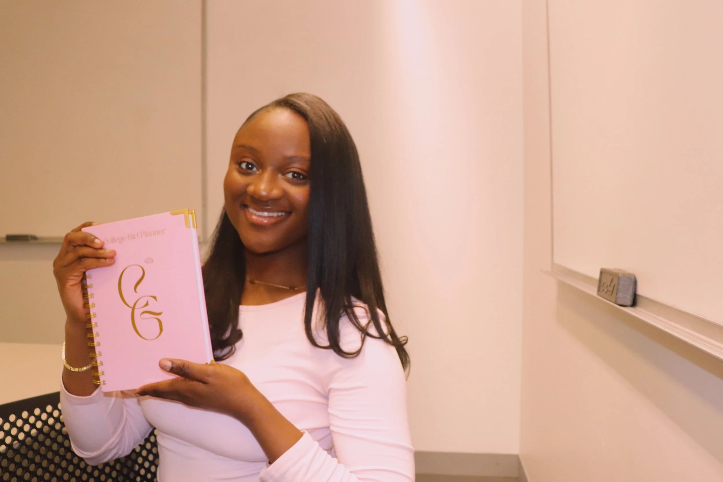 Young woman with long black hair smiling and holding a pink planner with gold accents, sitting in a classroom.