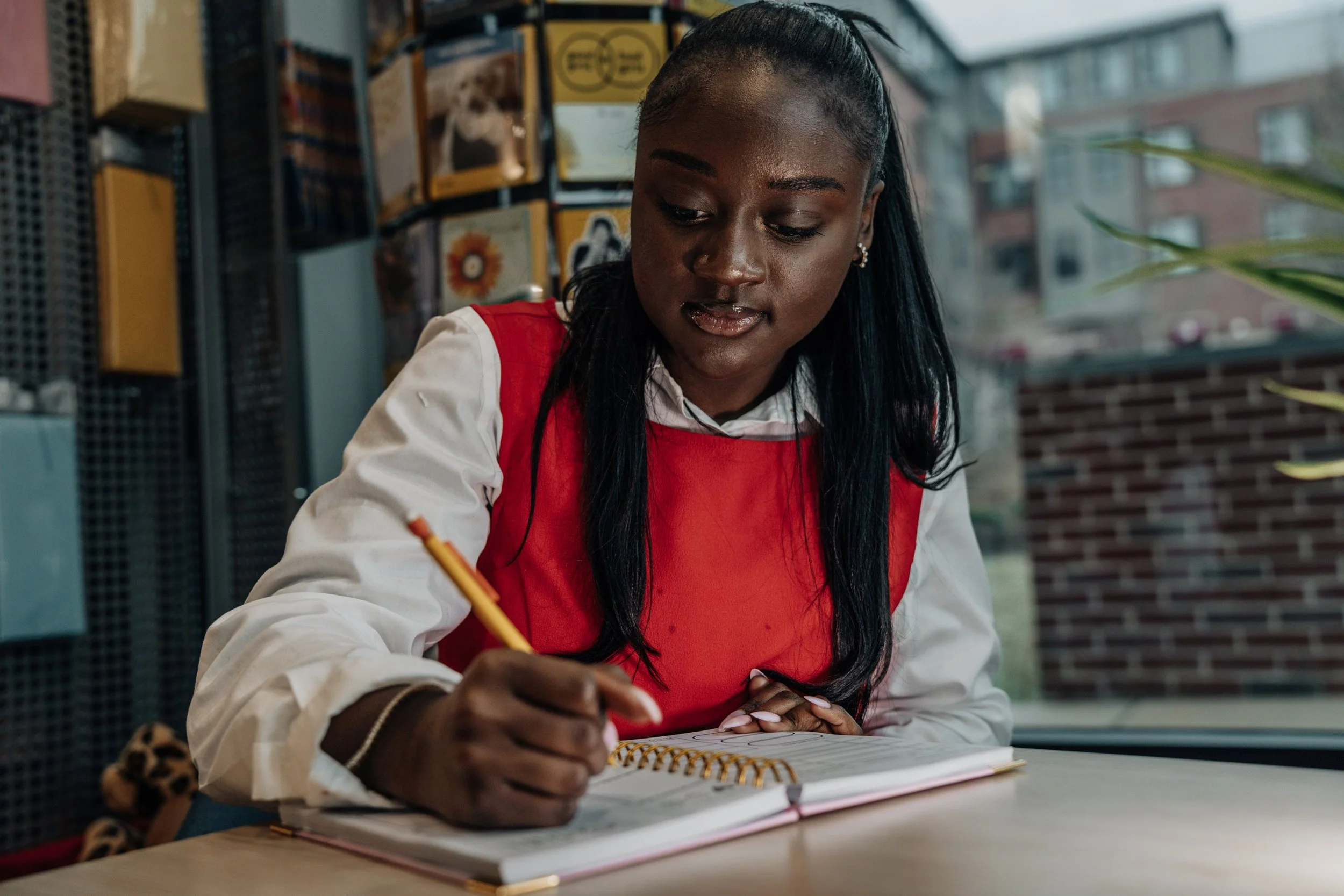 A young woman with dark skin and long black hair, wearing a red vest over a white shirt, is writing in a notebook with a pencil at a desk in an indoor setting with a window and colorful posters in the background.