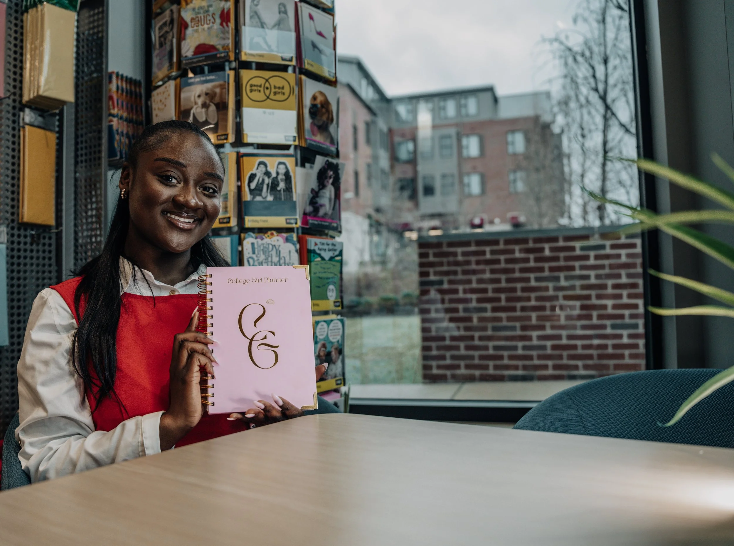 A smiling young woman sitting at a table inside a bookstore or library, holding a pink college girl planner with the initials 'CG' on the cover, with bookshelves and a window showing a brick building and trees outside.
