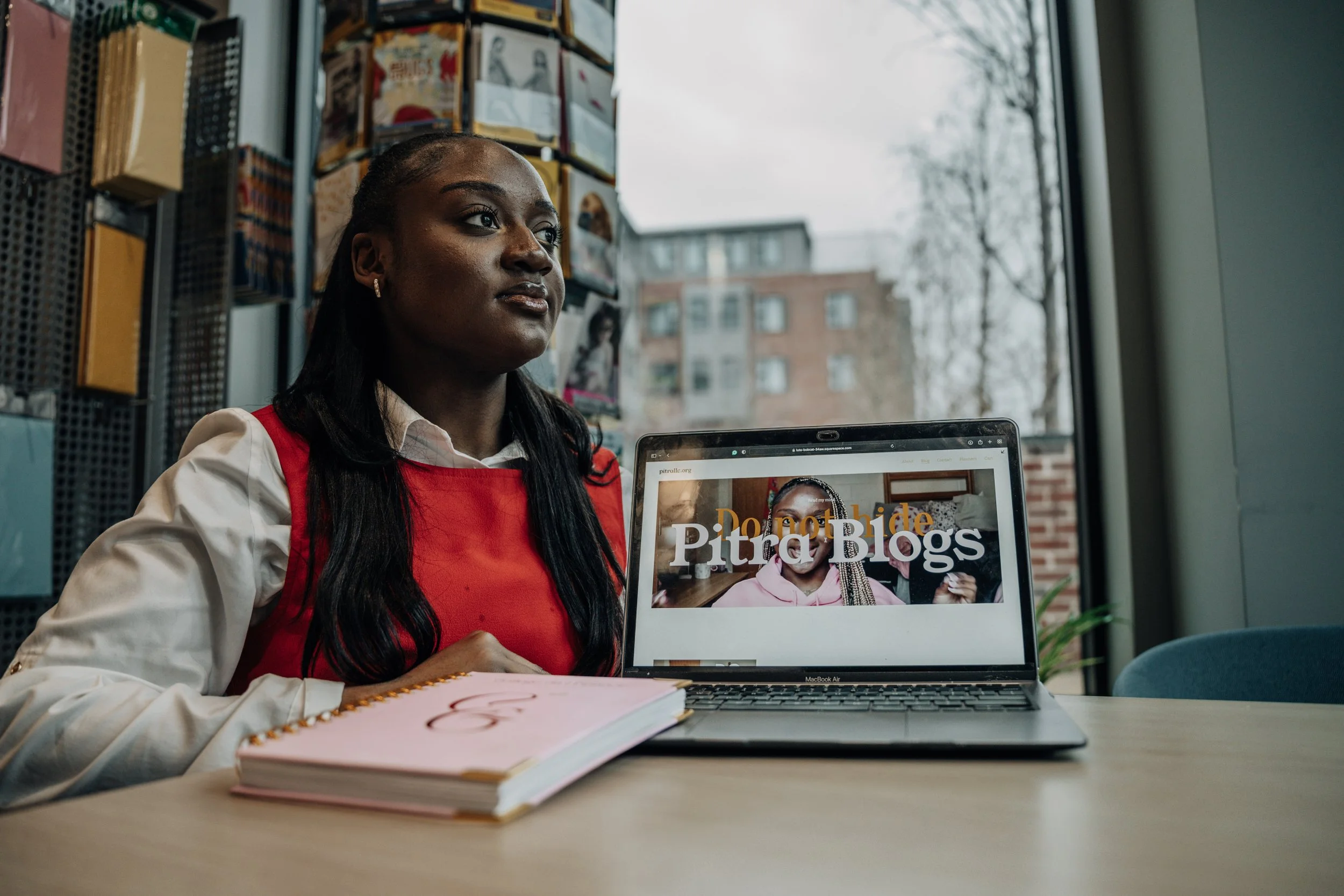 A woman wearing a white shirt and red sleeveless dress sitting at a table near a window, with a closed laptop displaying a blog titled "Pitra Blogs" and a pink notebook in front of her.