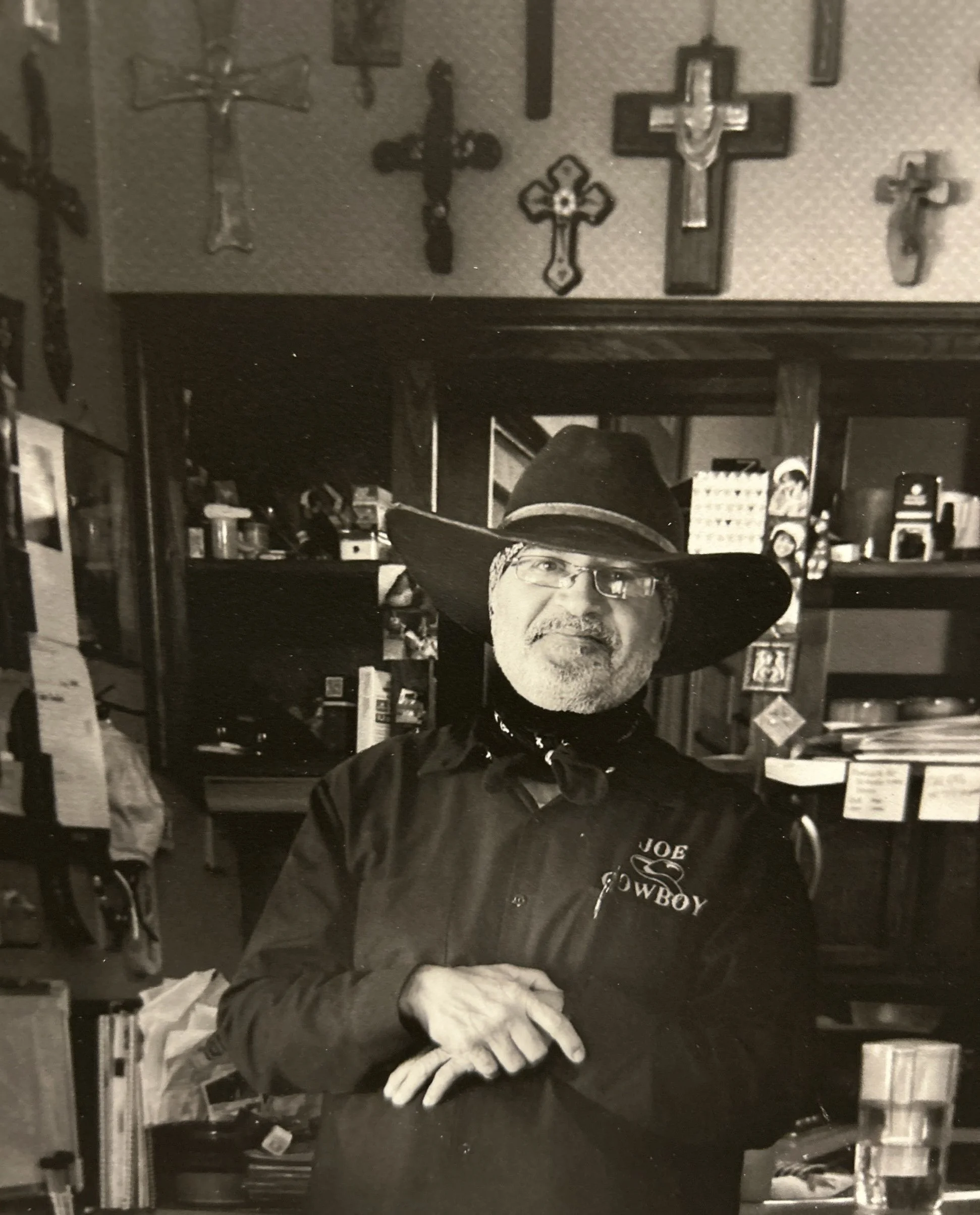 A man wearing glasses, a cowboy hat, and a dark shirt with 'Joe Cowboy' embroidered on it, standing behind a counter in a store with crossed hands. The store has shelves with various items and cross pictures on the wall.
