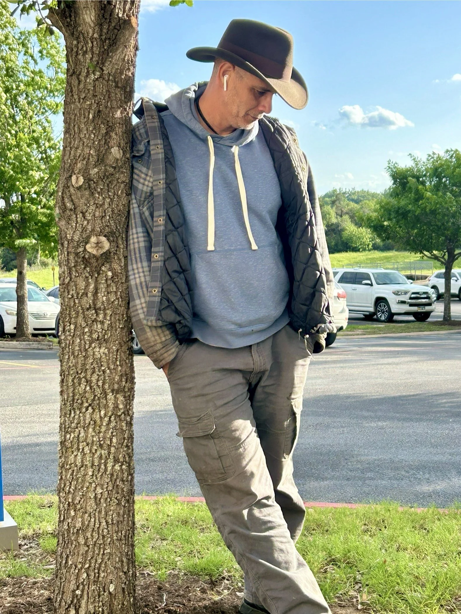 A man leaning against a tree in a parking lot, wearing a cowboy hat, gray hoodie, plaid shirt, and cargo pants, with AirPods in his ears and hands in his pockets.