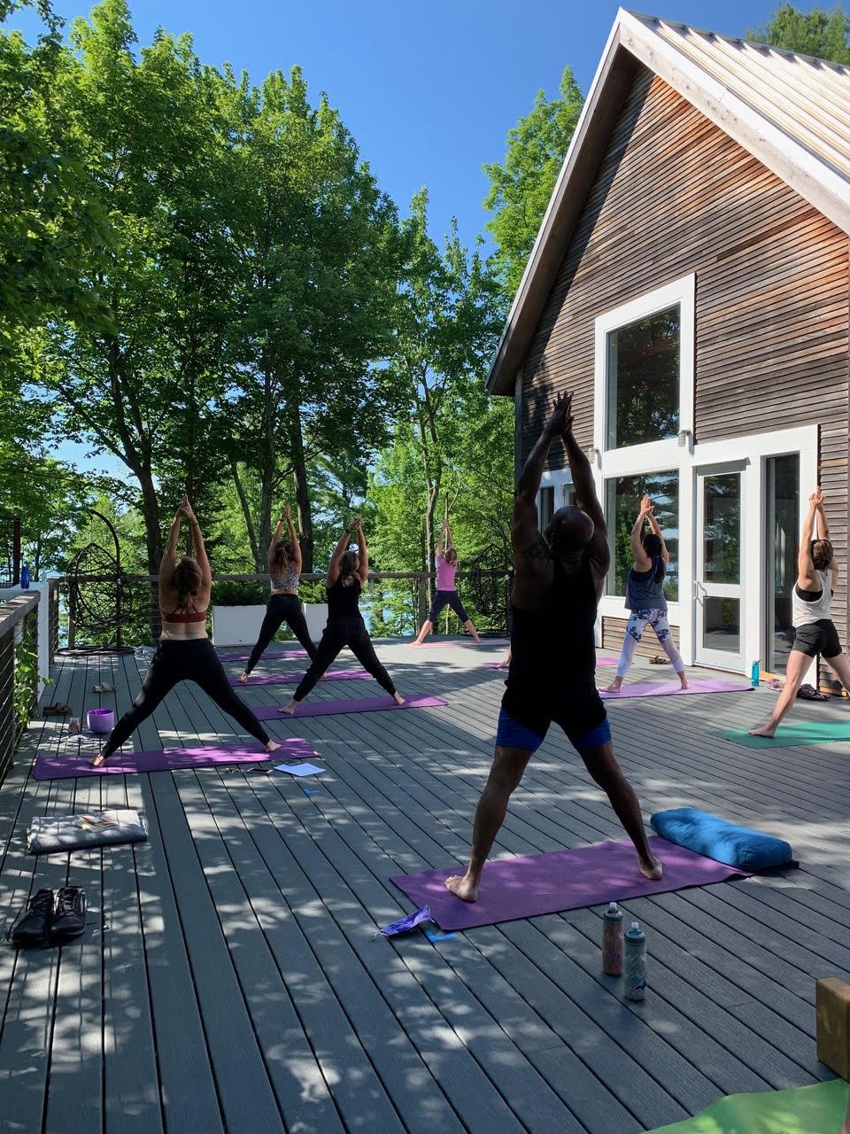 Group of people practicing yoga outdoors on a deck surrounded by trees, with a modern house in the background.