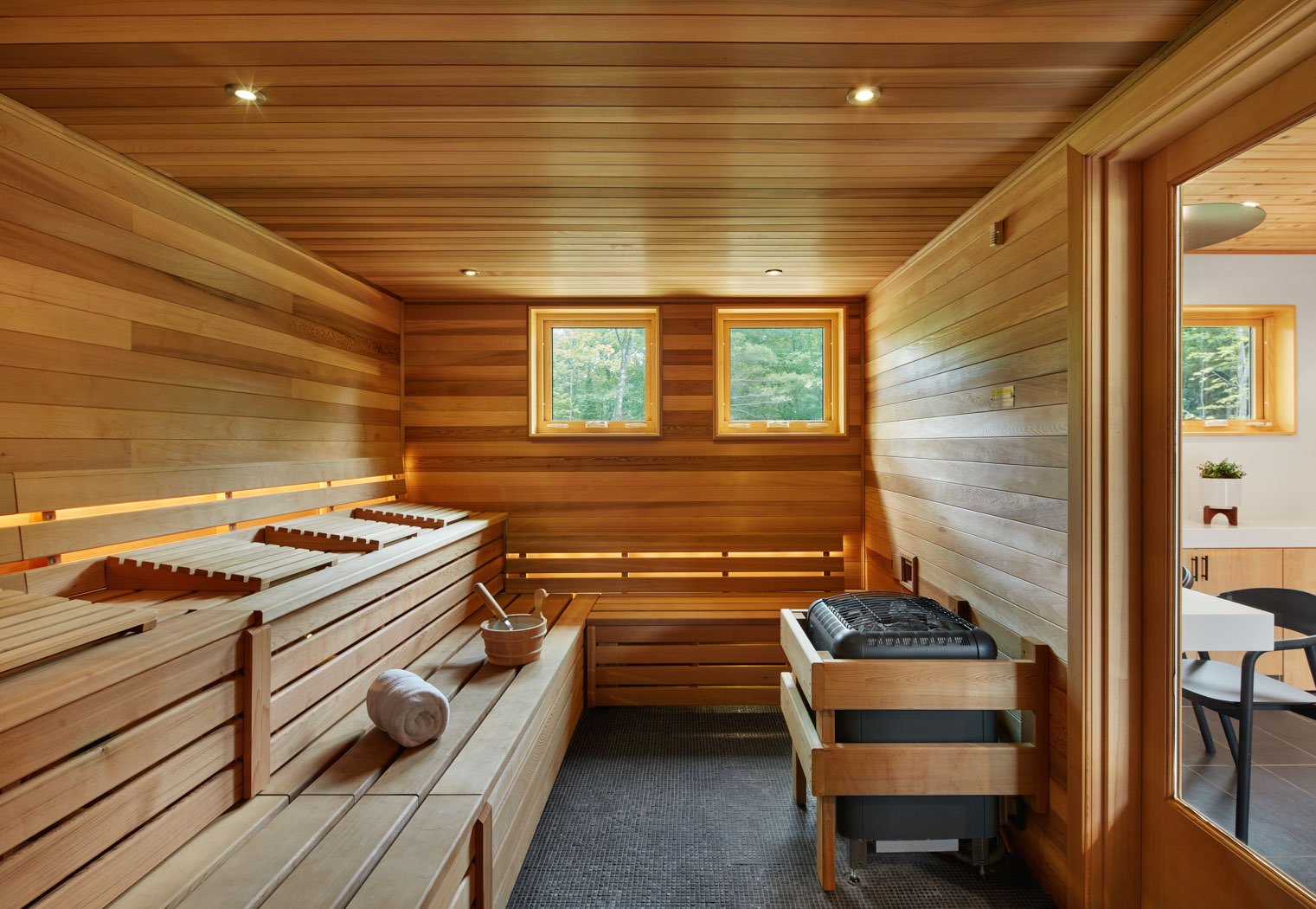 Wood-paneled sauna room with benches, a towel, a bucket, and a heater, with two small windows showing greenery outside.