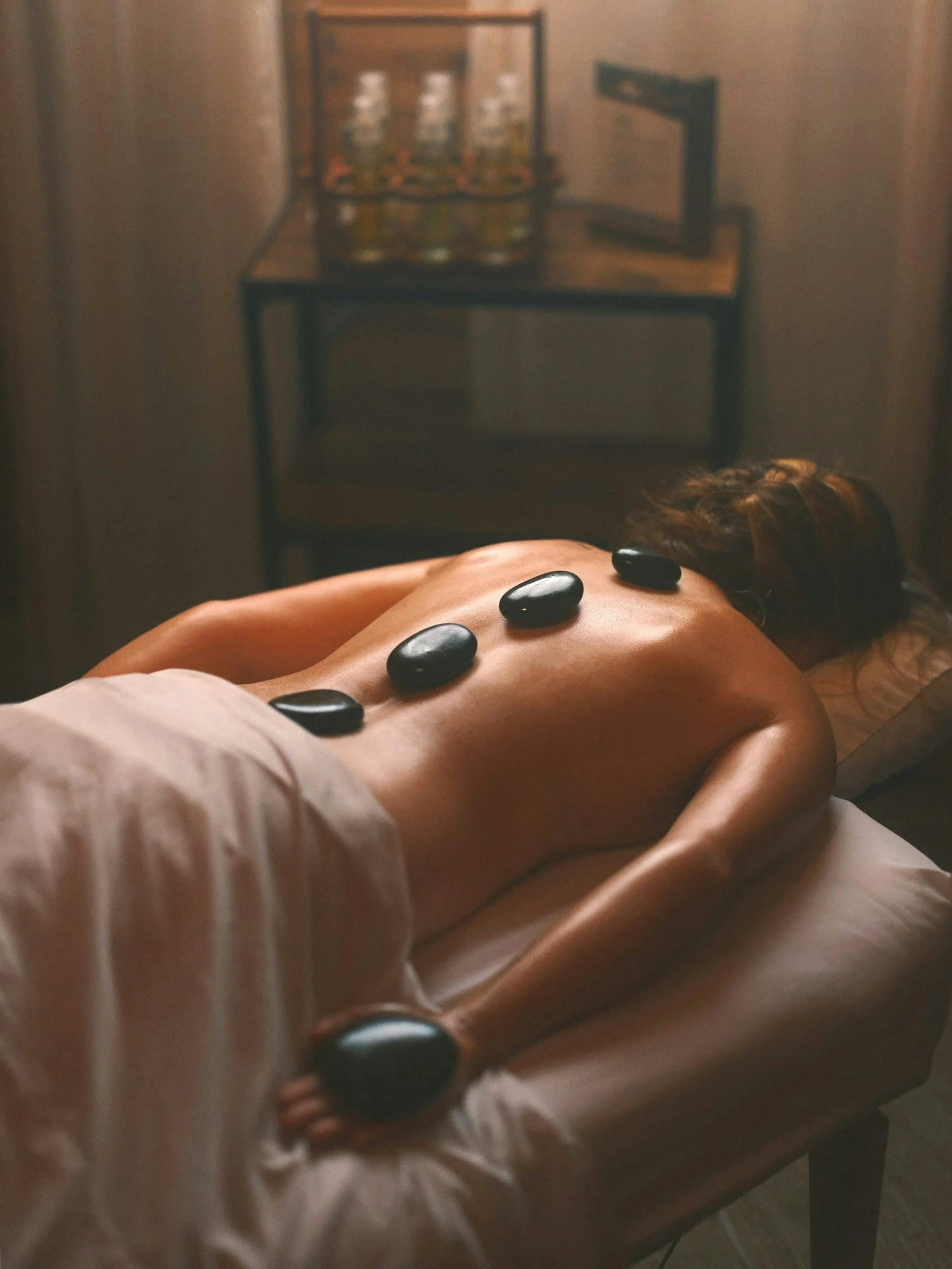 A person lying face down on a massage table with black stones placed along their back for hot stone massage, in a dimly lit room.
