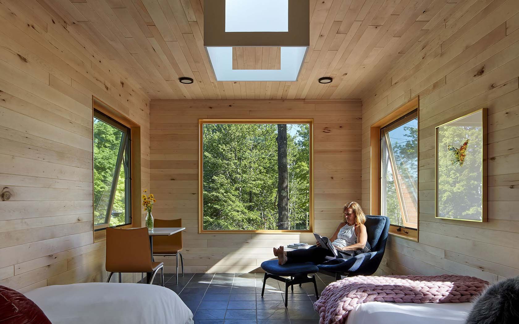 A woman sitting in a blue armchair reading a book or tablet in a room with light wooden walls and large windows showing trees outside, with a bed and a small table with flowers.