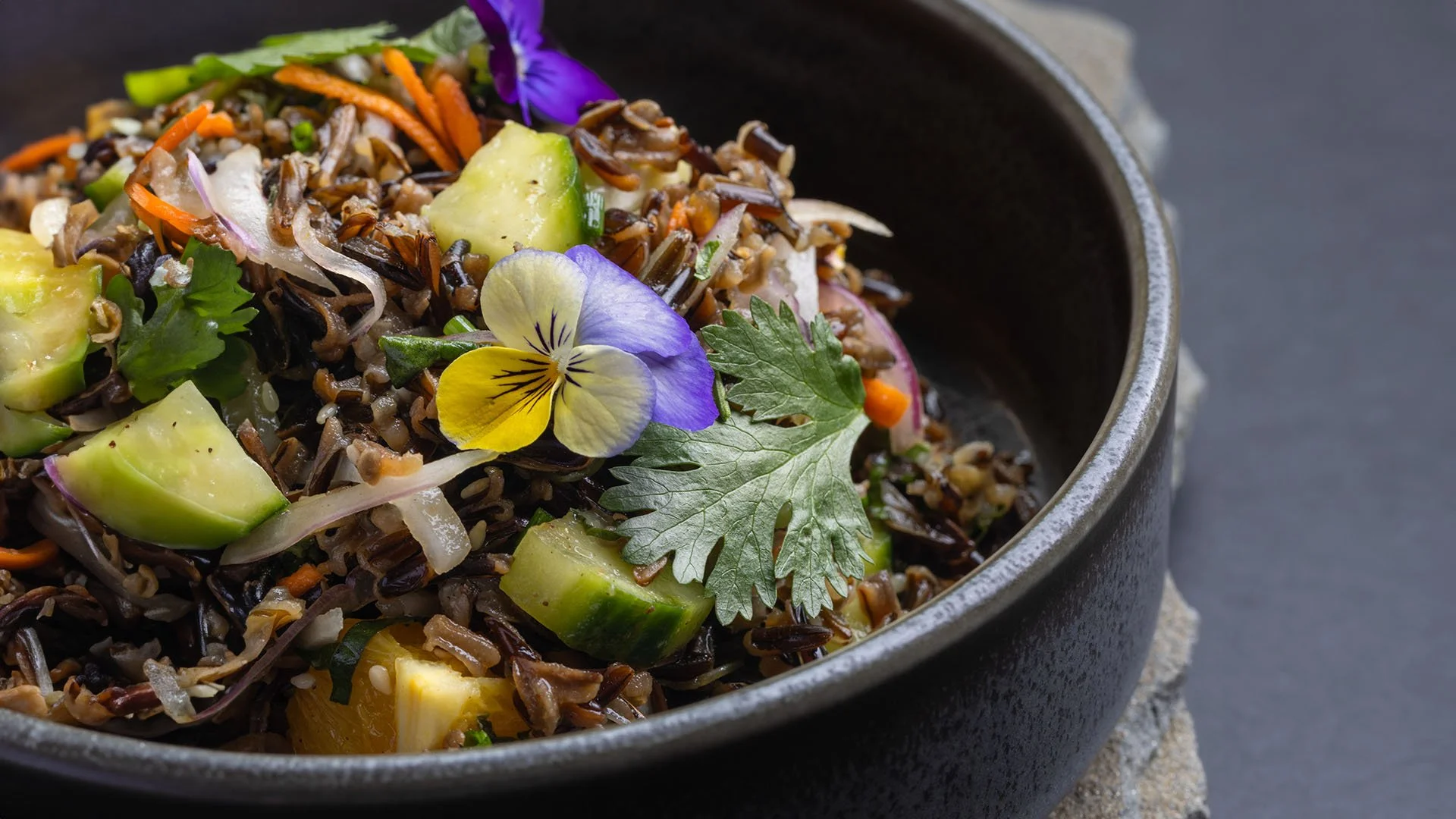 A black bowl of cooked black rice salad garnished with slices of cucumber, chopped red onion, fresh cilantro leaves, and edible purple and yellow flowers on top.