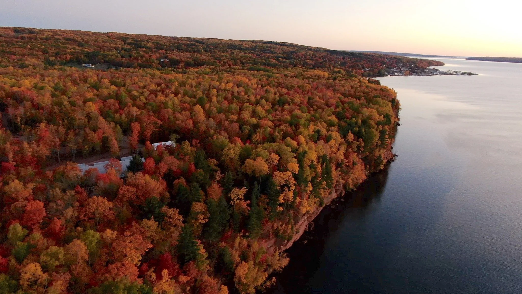 Aerial view of a river bordered by a forest with colorful autumn foliage, and a small town in the distance at sunset.