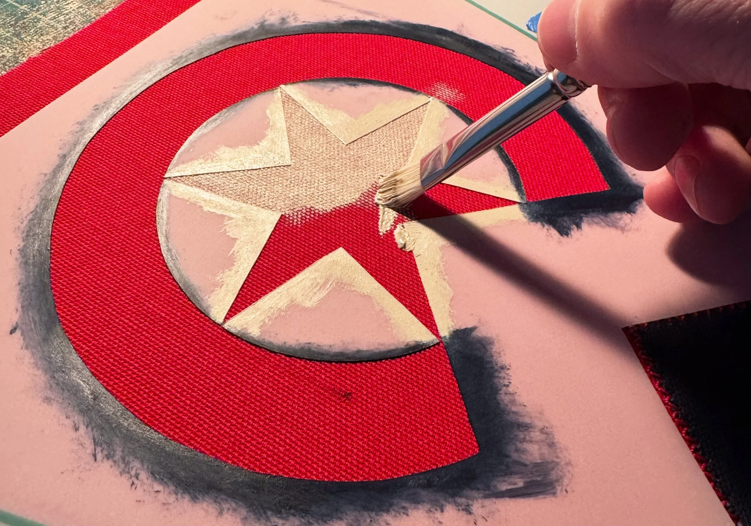A person is painting a gold star inside a red circle with a black outline on pink paper, using a silver paintbrush.