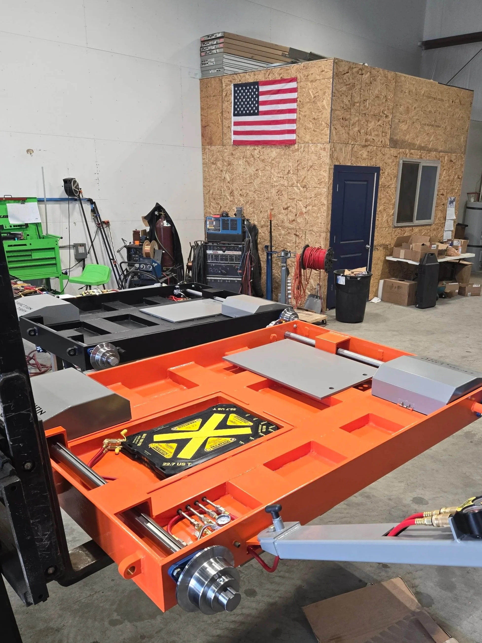 A construction or workshop space with a partially assembled piece of equipment or machinery. The foreground features an orange metal frame with various components and tools, including a black plate with a yellow warning sticker. In the background, there is a plywood structure with a small window and a door, a U.S. flag hanging on the plywood, a stack of metal and wood boards on top of the structure, and various workshop tools and materials scattered around.