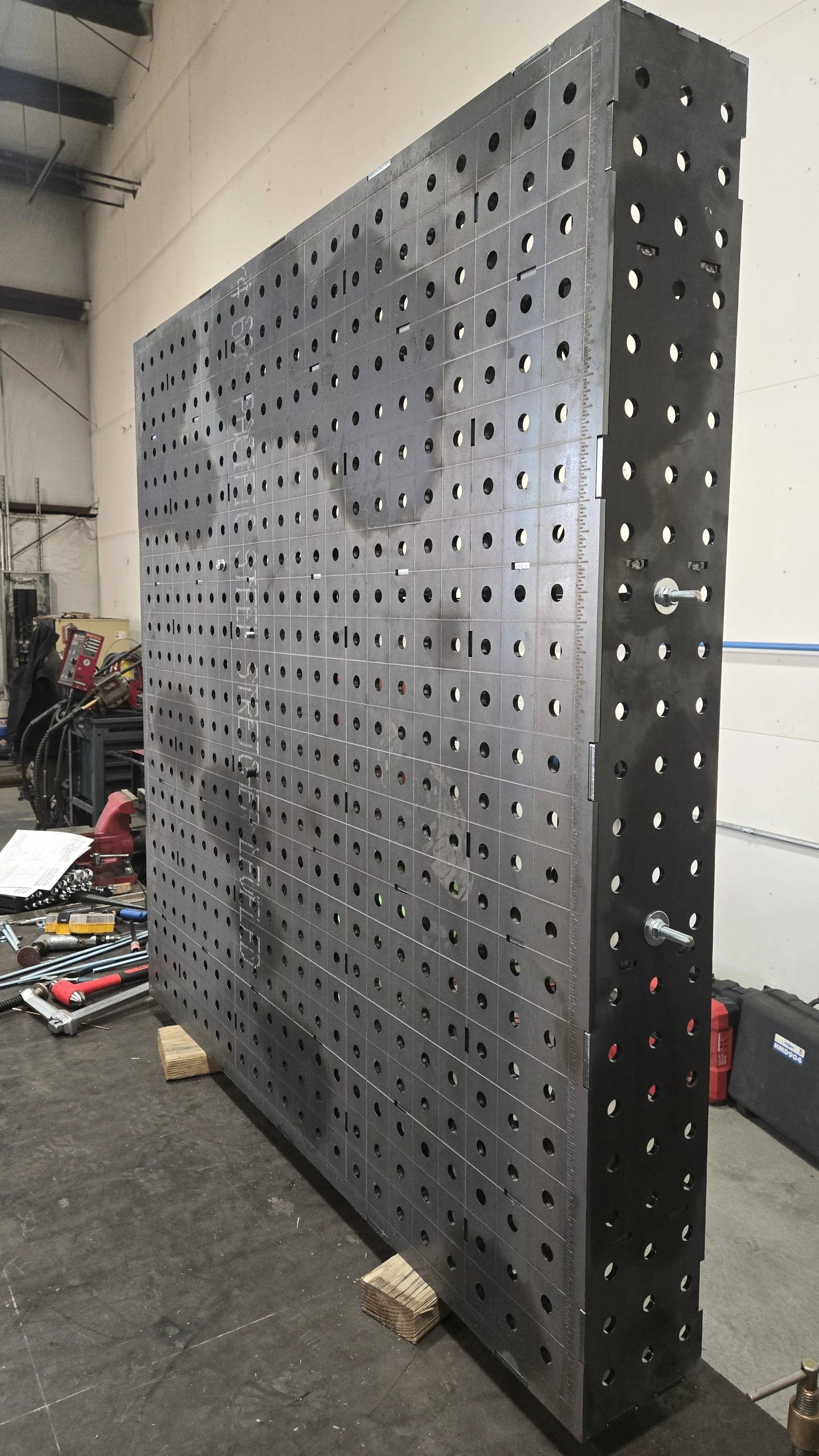 Large metal assembly with multiple evenly spaced holes, standing upright in an industrial workspace. Various tools and equipment are scattered on the workbench nearby.