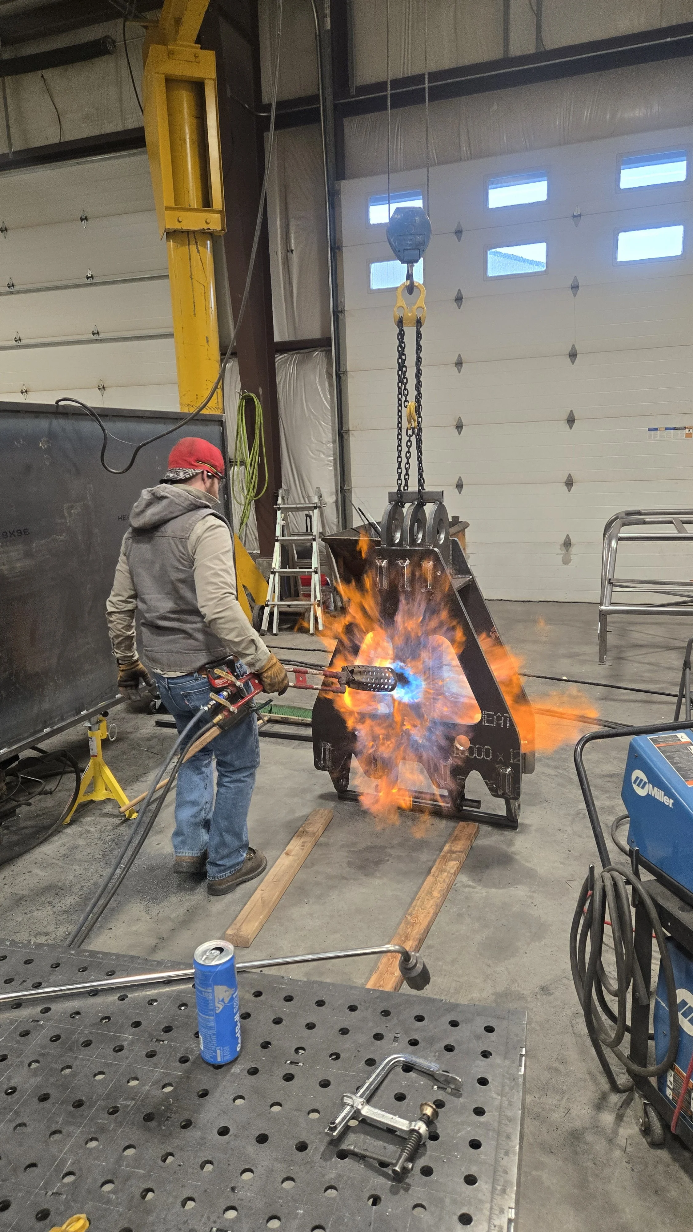 A person wearing a red head covering, glasses, a gray vest, and jeans is using a blowtorch to cut a large metal piece in an industrial workshop. Sparks and flames are visible from the cutting process, with safety equipment and tools around.