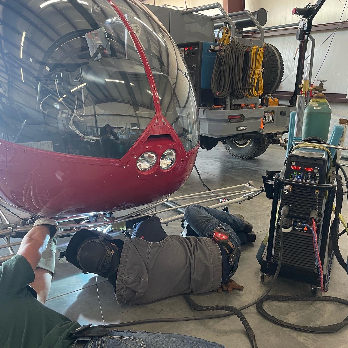 Two technicians are working underneath a red aircraft cockpit or canopy on the ground in a garage or hangar, with one lying on their back and the other kneeling. There is a welding or air conditioning machine with cables nearby. In the background, a utility truck with coiled hoses and a spare tire is parked beside a wall.