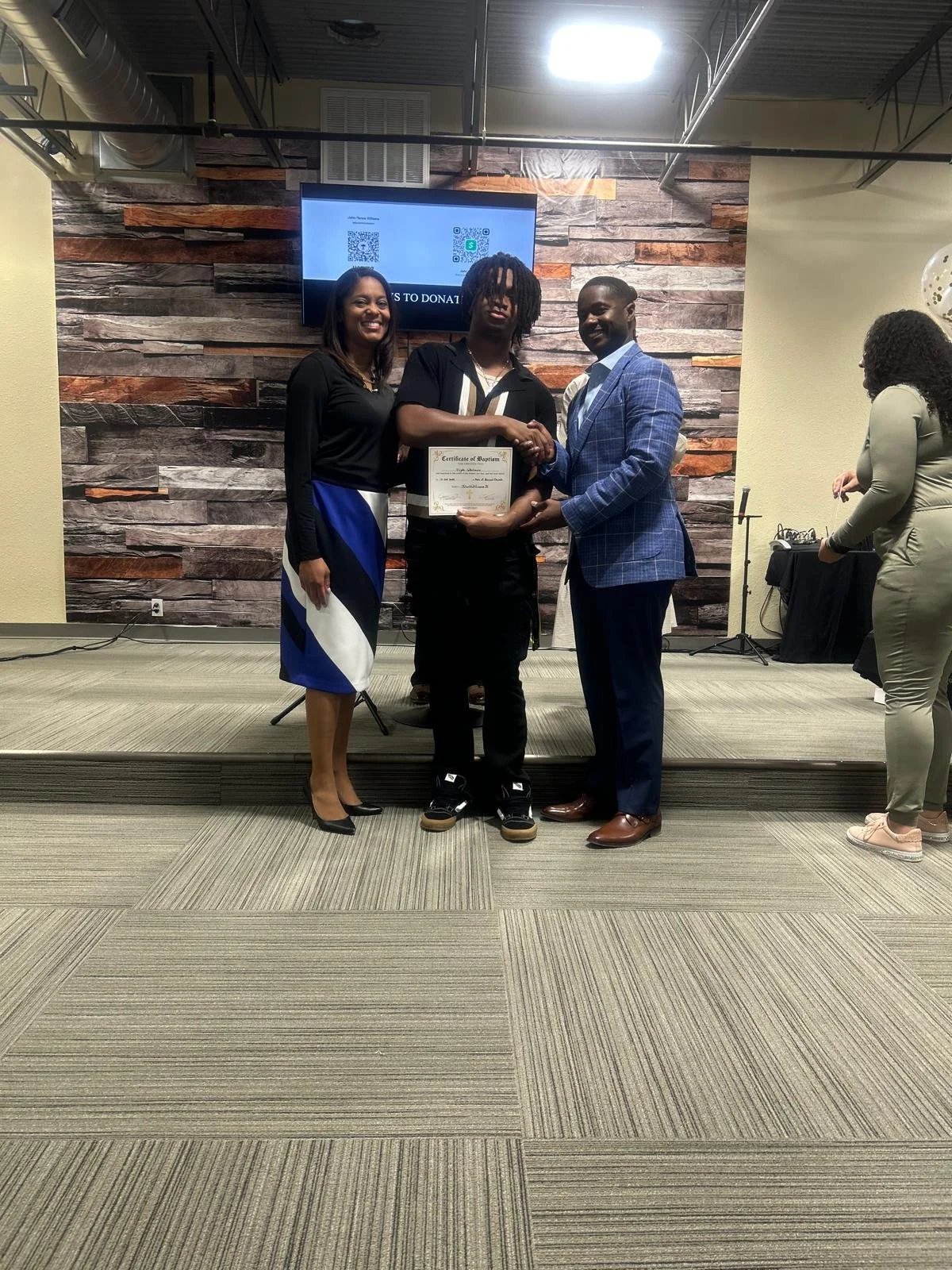 Three people standing on a stage, with a man in the middle handing a certificate to a young man. Two women are standing next to the young man, smiling. The background features a wood-paneled wall and a screen displaying QR codes, with another woman and equipment to the side.
