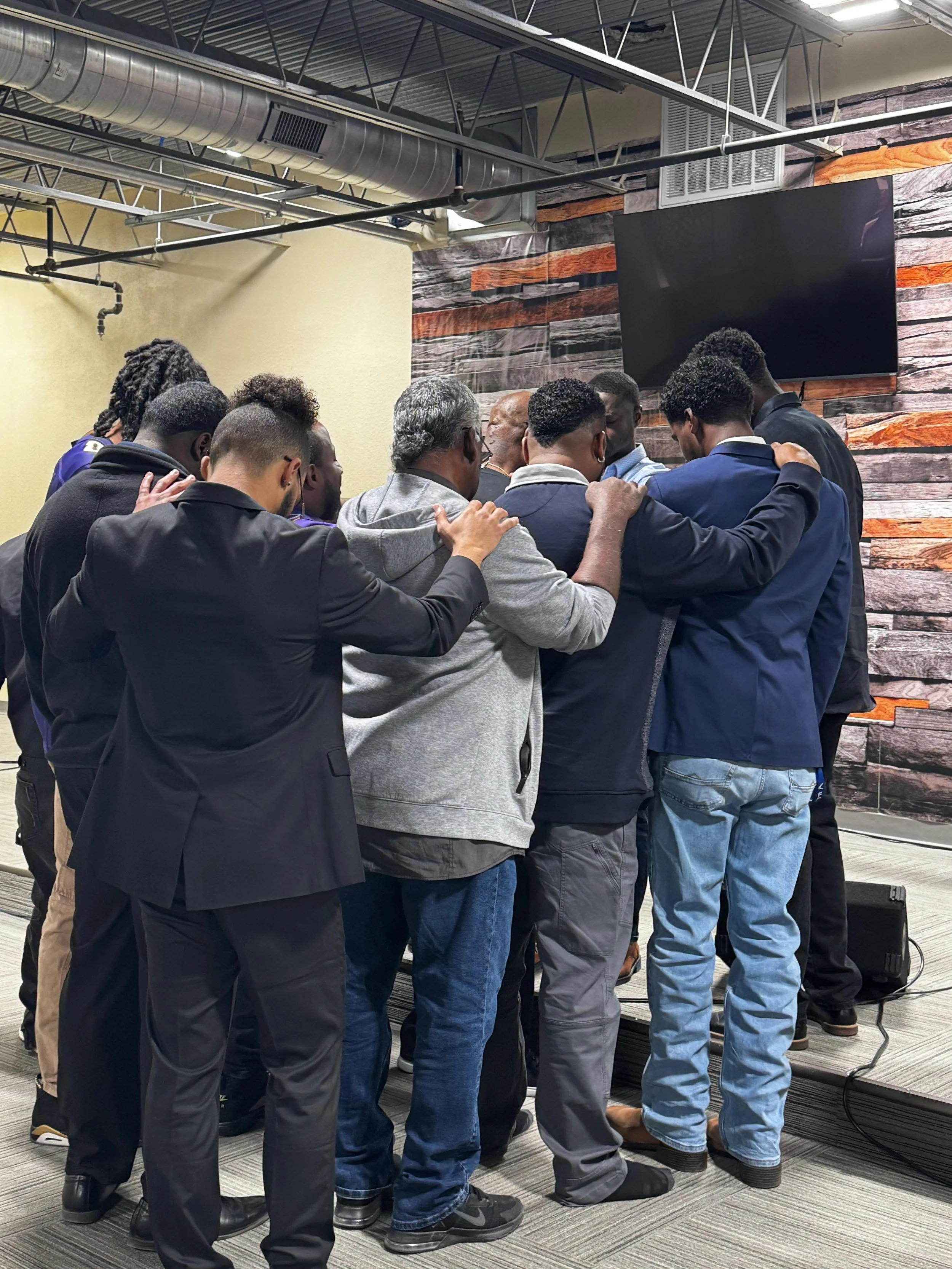 A group of people with their arms around each other's shoulders, standing close together with heads bowed, in a room with a wood-paneled accent wall and a television screen mounted on the wall.
