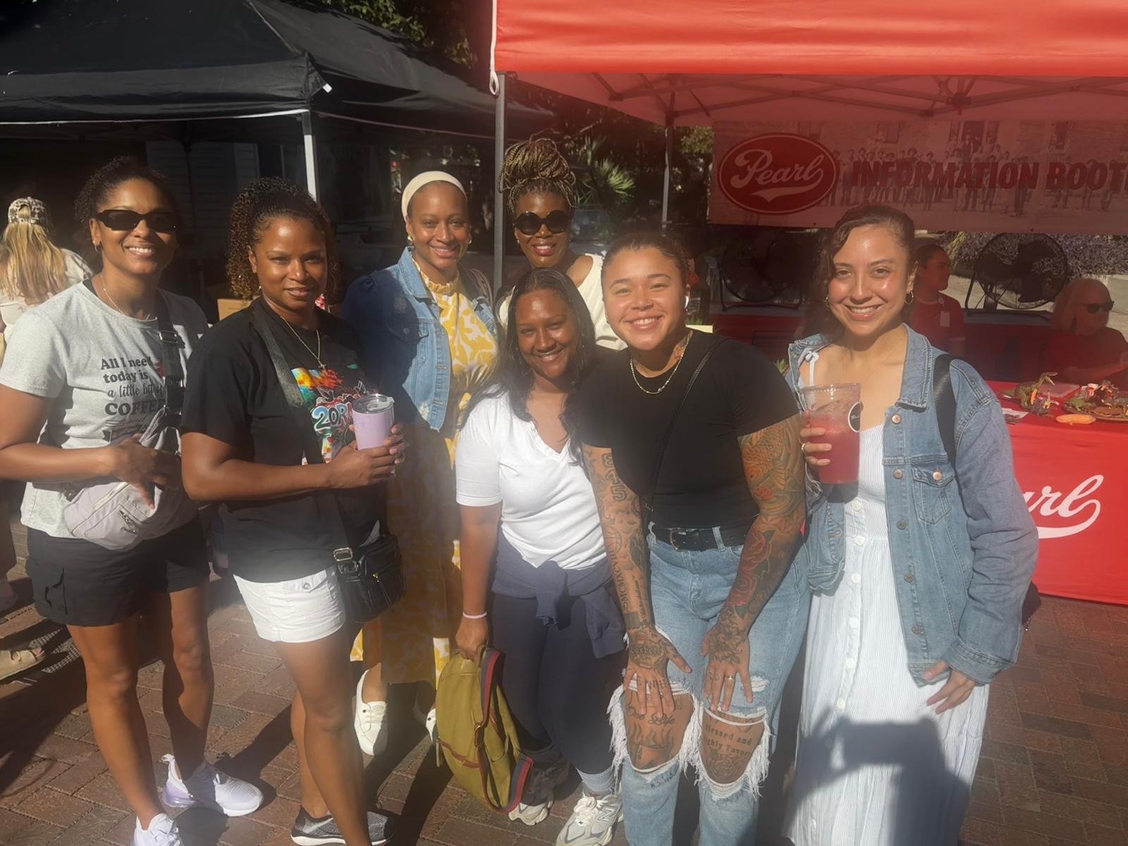 Group of seven women smiling at an outdoor event, standing in front of a red tent with a banner, some holding drinks, dressed casually.