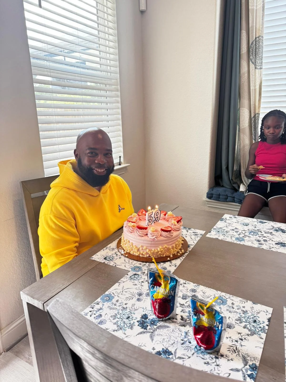 A man in a yellow hoodie celebrating his 59th birthday with a pink birthday cake with strawberries and candles, two Capri Sun drinks, and a young girl sitting in the background holding a snack.