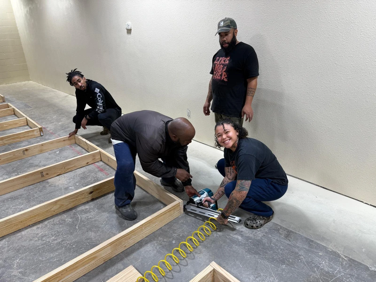 Four people working together to build a wooden structure inside a room. One person is using a power tool, and the others are assisting or observing.