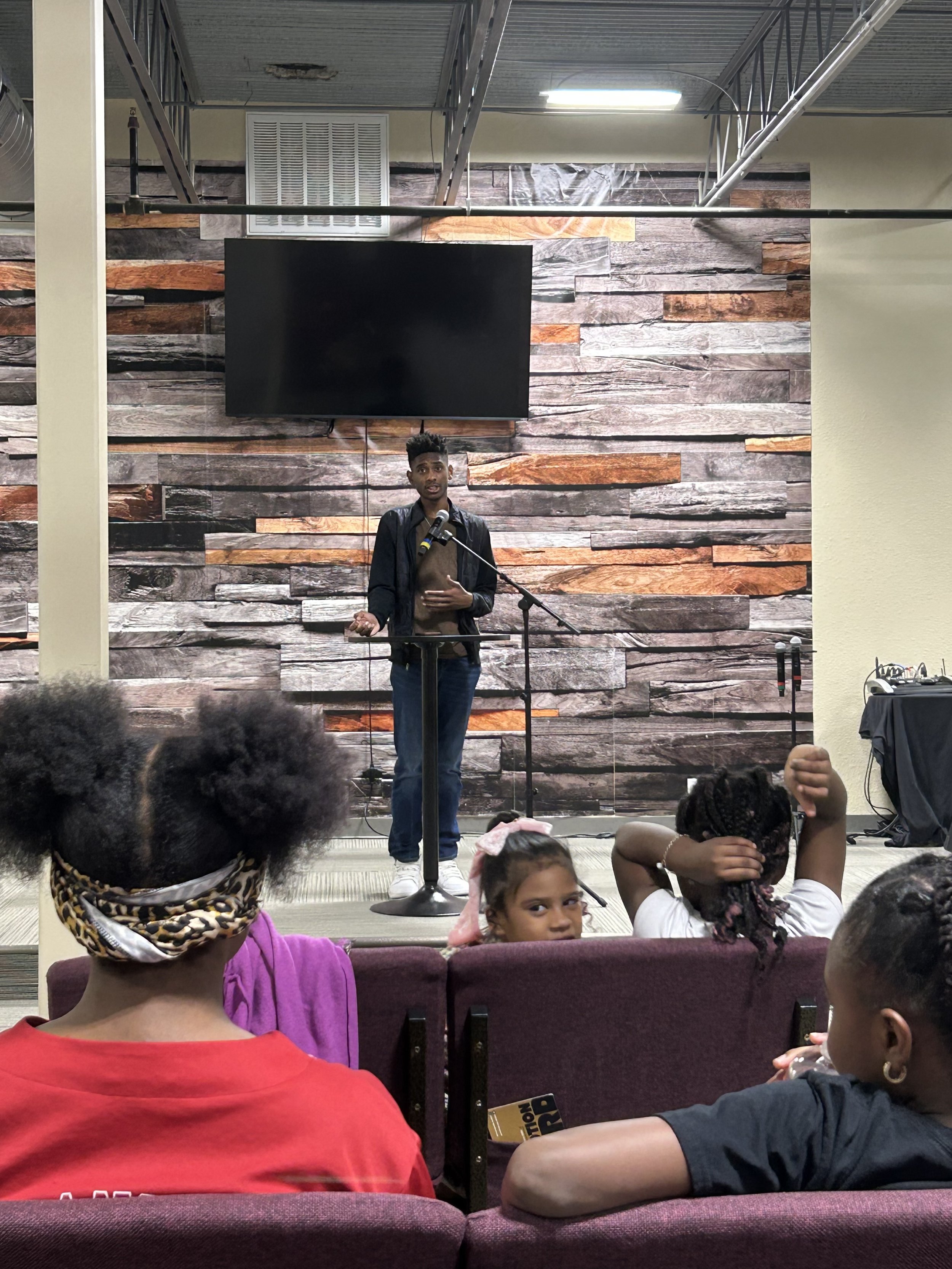 A young person stands at a microphone on a stage, speaking to an audience. The stage backdrop is made of textured wood panels, and there is a large TV screen above them. In the foreground, several children are seated, watching the speaker.