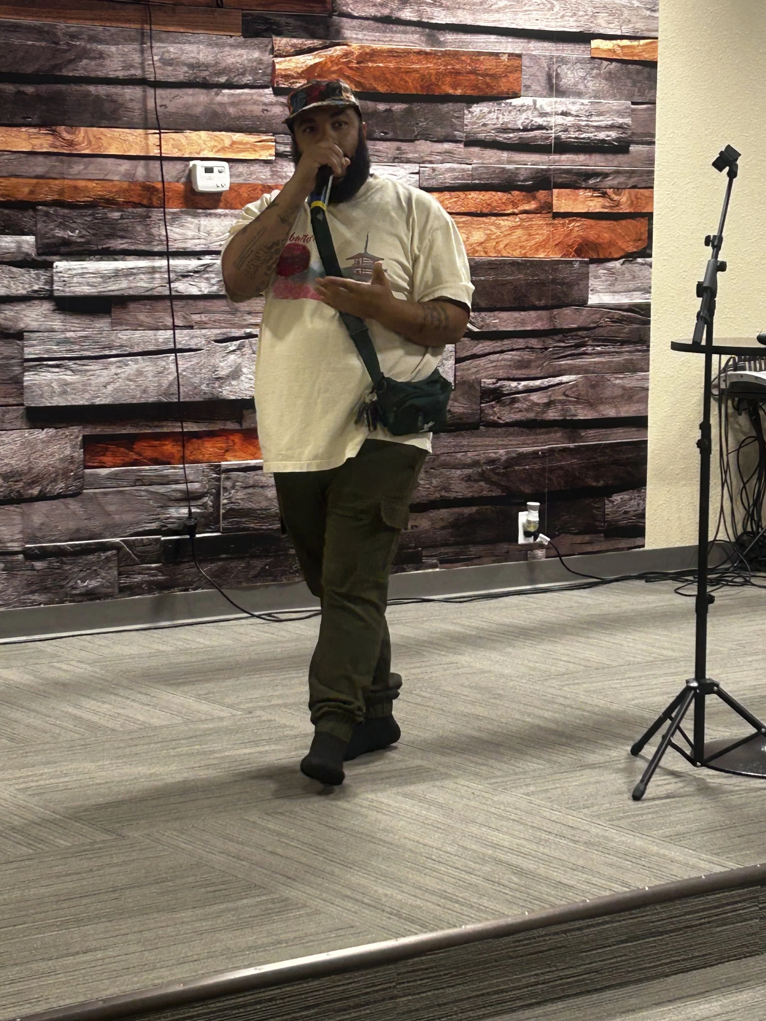 Man performing with a microphone against a wooden backdrop, wearing a white t-shirt and camouflage hat.