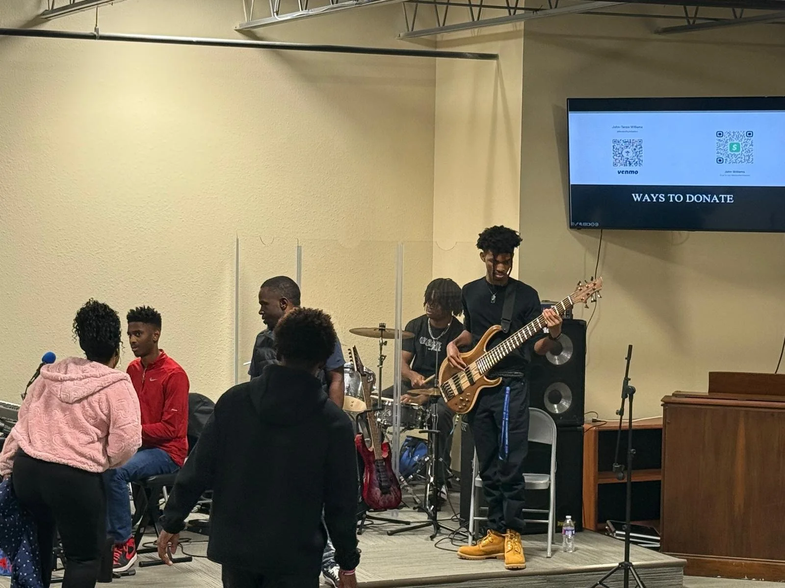 A band performing on stage with three members playing instruments including bass guitar and drums, and three people in the audience engaged in conversation, with a large screen displaying donation instructions in the background.