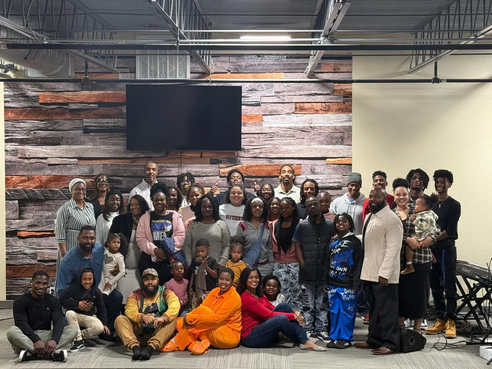 Large group of people posing indoors in front of a decorative wood-patterned wall. The group includes adults and children, with some people seated on the floor and others standing. There's a digital piano to the side.