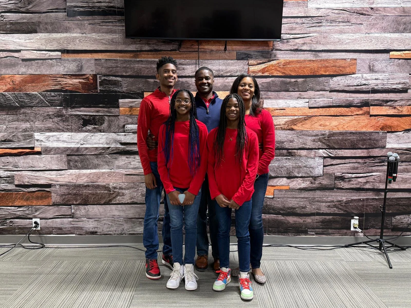 Group of five people standing together in front of a decorative wood-paneled wall, smiling, with a television mounted above them and a microphone stand to the right.