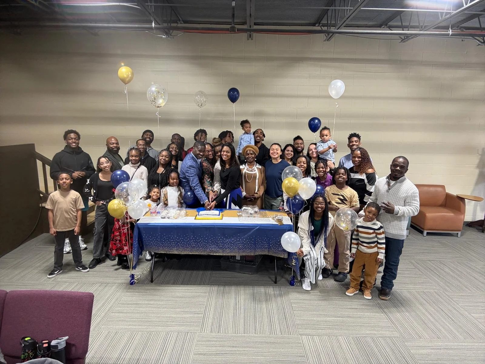 A large group of people gathered for a celebration, with a table decorated with a blue tablecloth, balloons, and a cake, in a spacious indoor room with group photos and smiling faces.