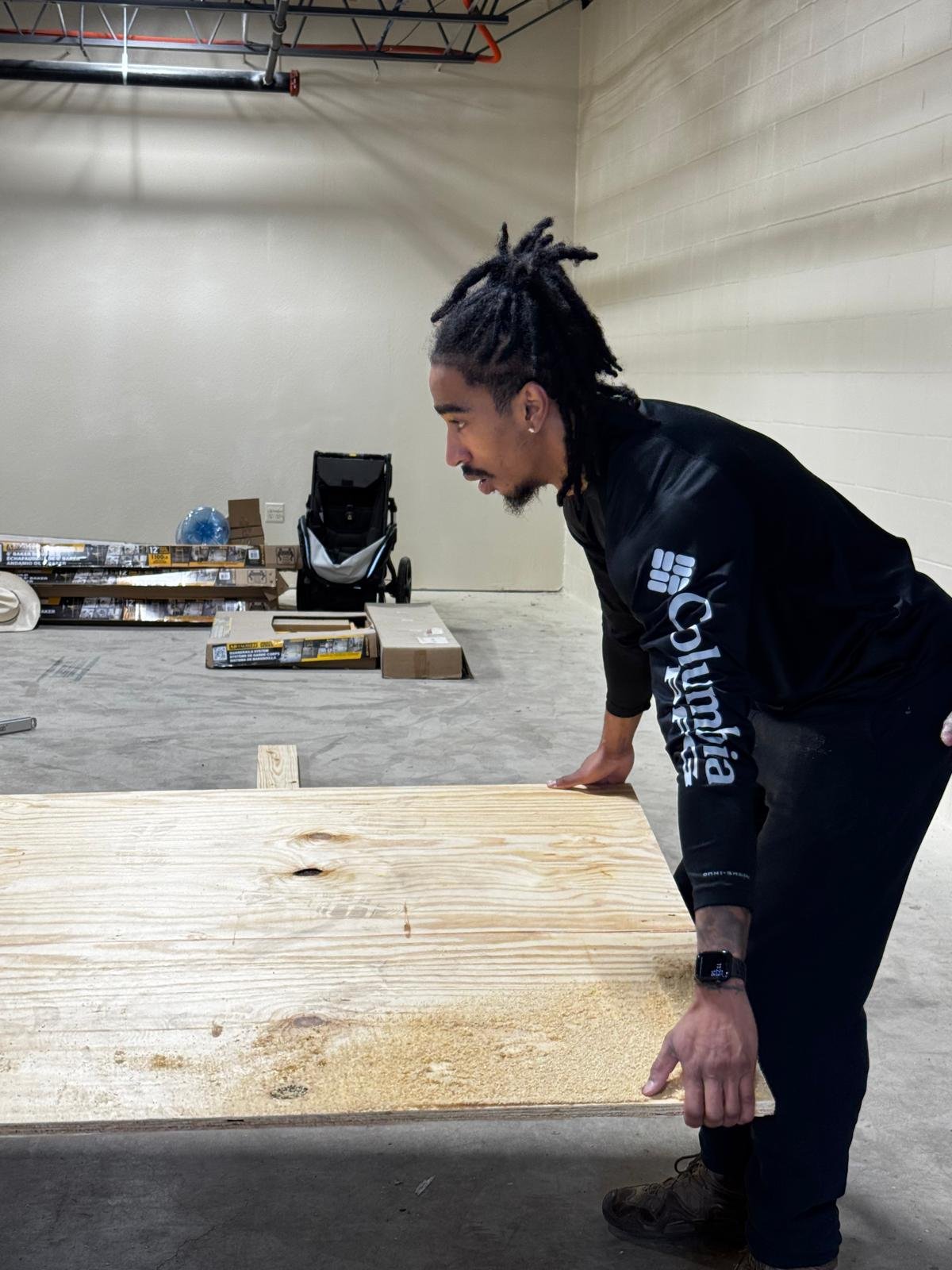 A man with dreadlocks wearing a black Columbia jacket is bending over a large wooden board, holding onto its edges in an indoor workspace with tools and boxes in the background.