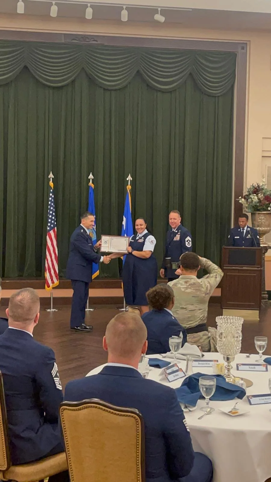A military ceremony where a female officer is being awarded a certificate by a male officer, with other officers standing behind her, in a formal setting with green curtains and flags, and an audience seated at tables.