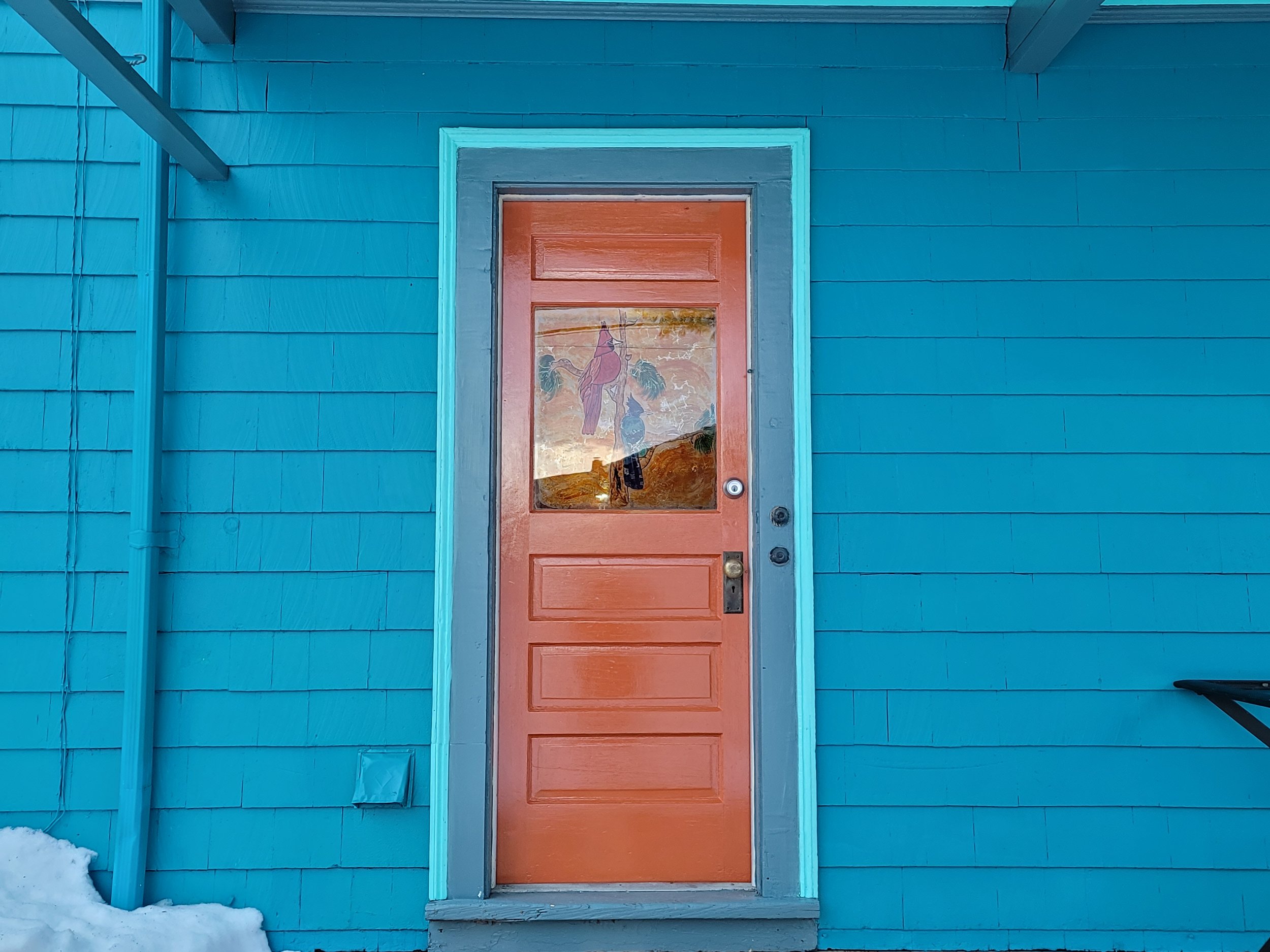 High gloss orange porch door on a colonial in Dorchester, with a decorative window, set in a blue house with horizontal siding, with snow on the ground.