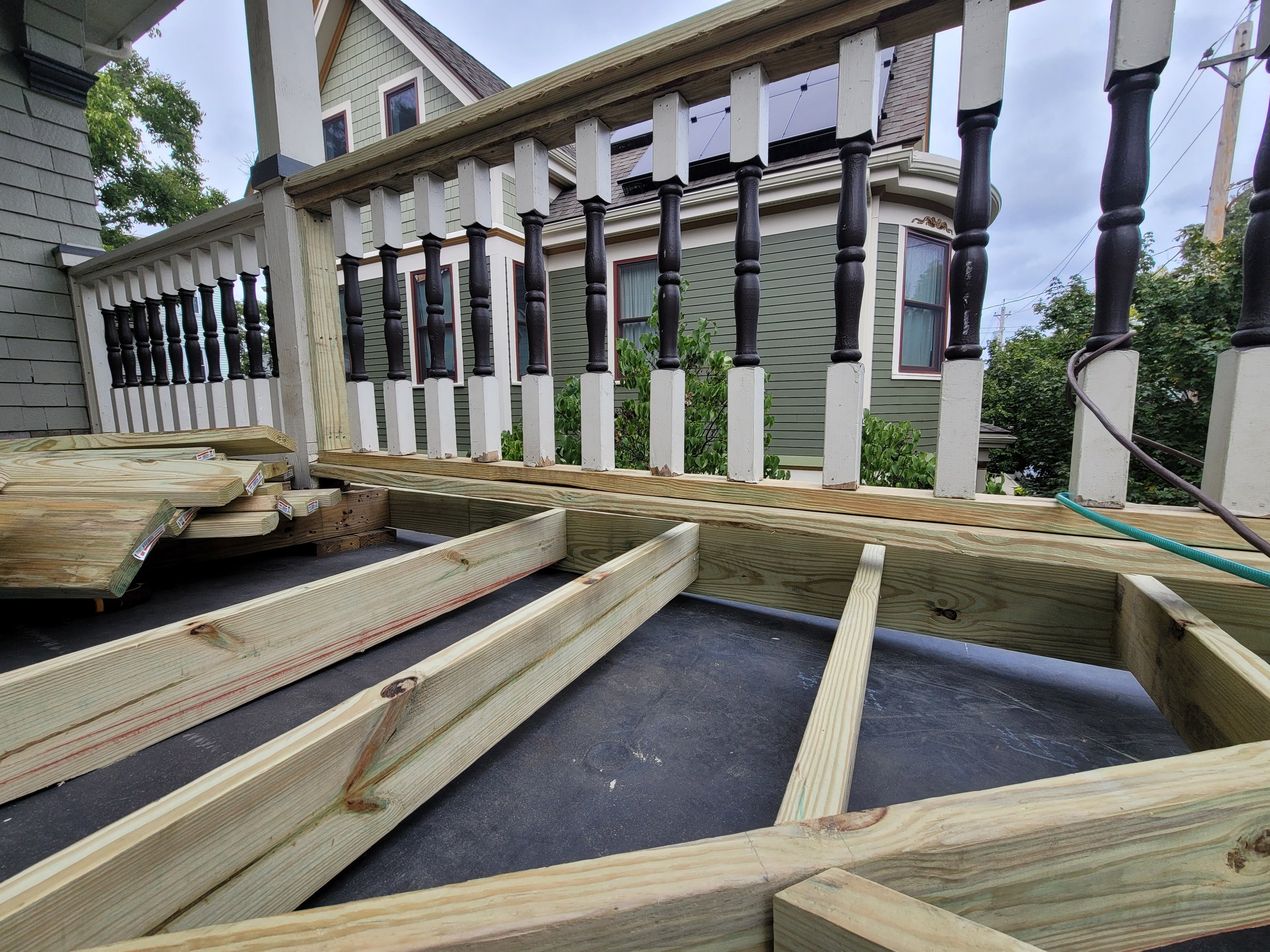 A progress picture of a deck repair and staining project in Dorchester, Boston where there is a partially constructed wooden deck with framing and railing in front of a house with green siding and white trim.