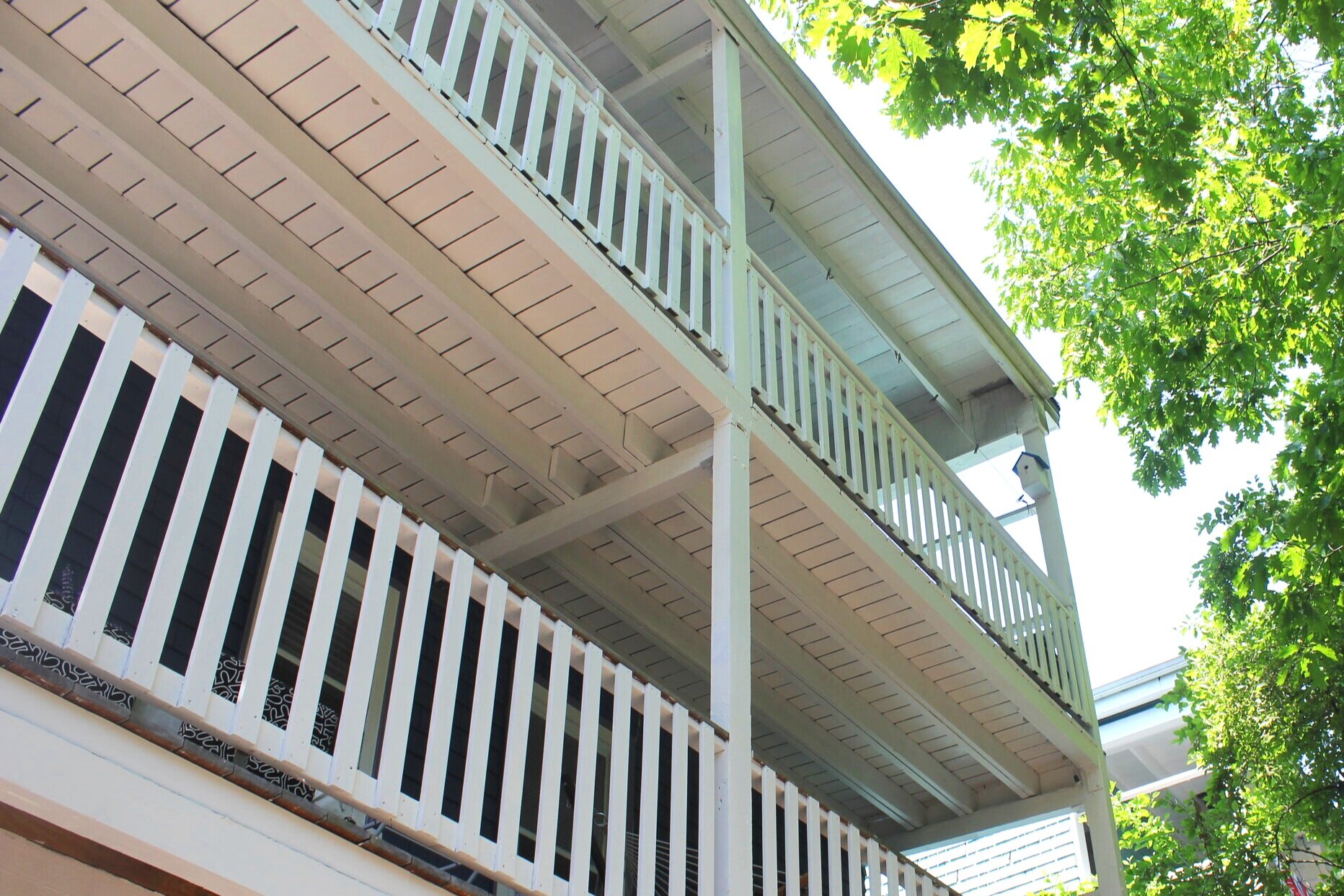 Multiple beige balconies with white railings on a Boston triple-decker building, surrounded by green tree leaves.