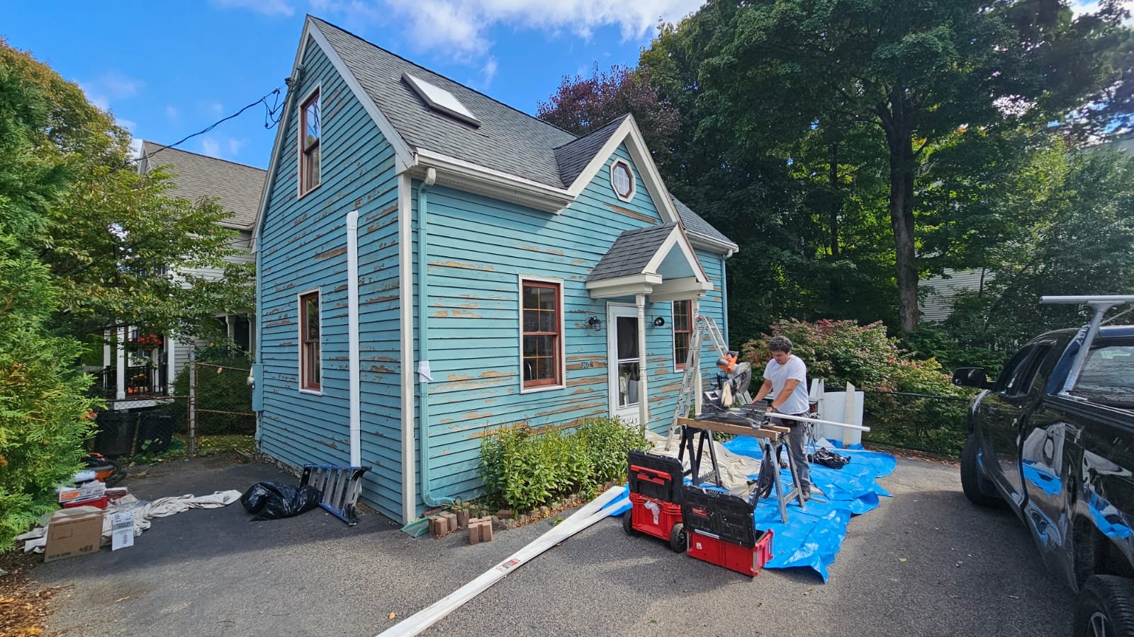 Two crew members from Ryan Painting carrying out a carpentry and restoration, exterior painting project on a blue, Roslindale cape-style house with brown window frames, surrounded by trees and parked cars, with tools and materials on the driveway.