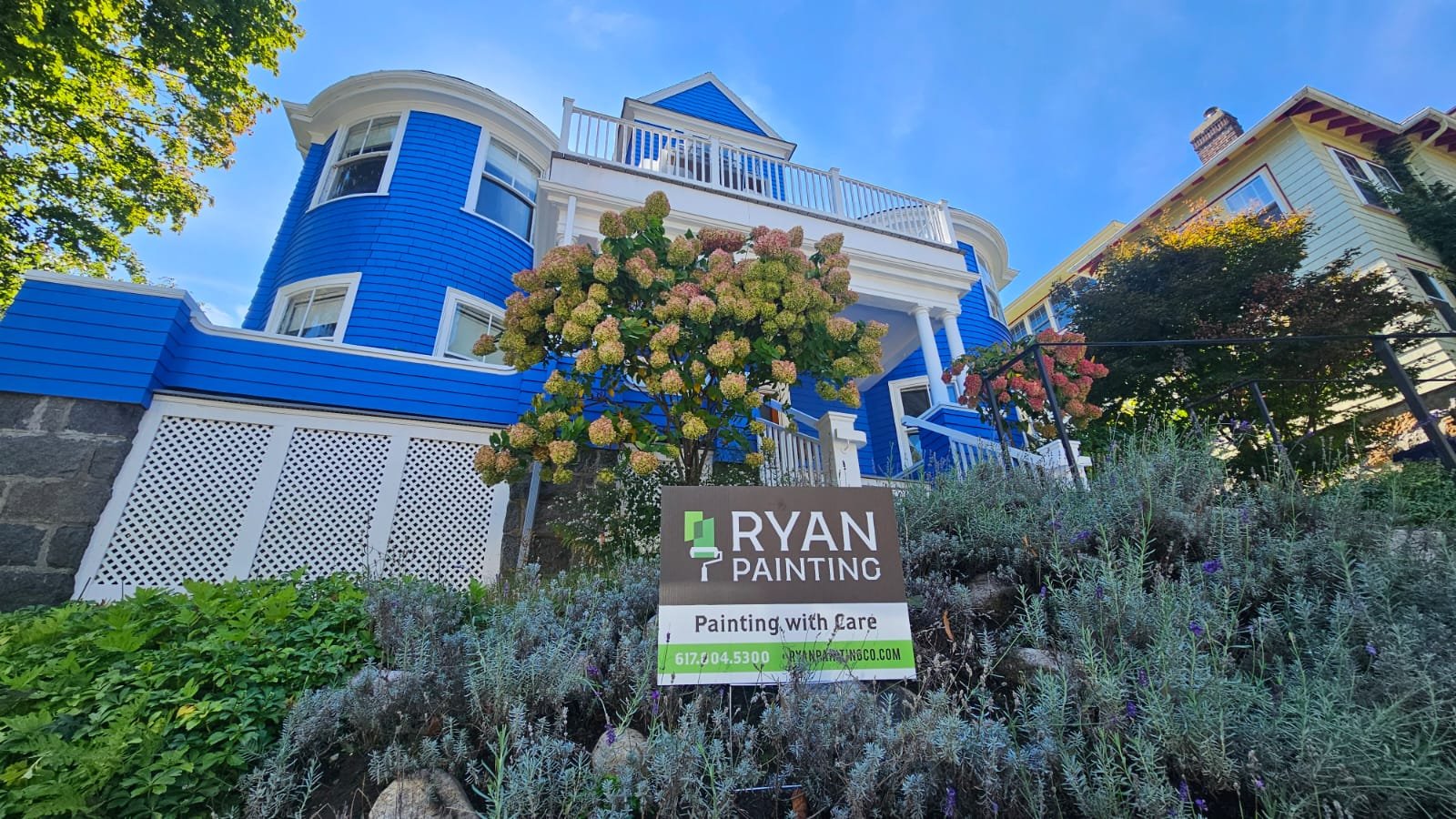 A colorful blue house on Hampstead Road, Jamaica Plain with white trim and a front porch, surrounded by plants and trees, with a sign for Ryan Painting in the front yard.