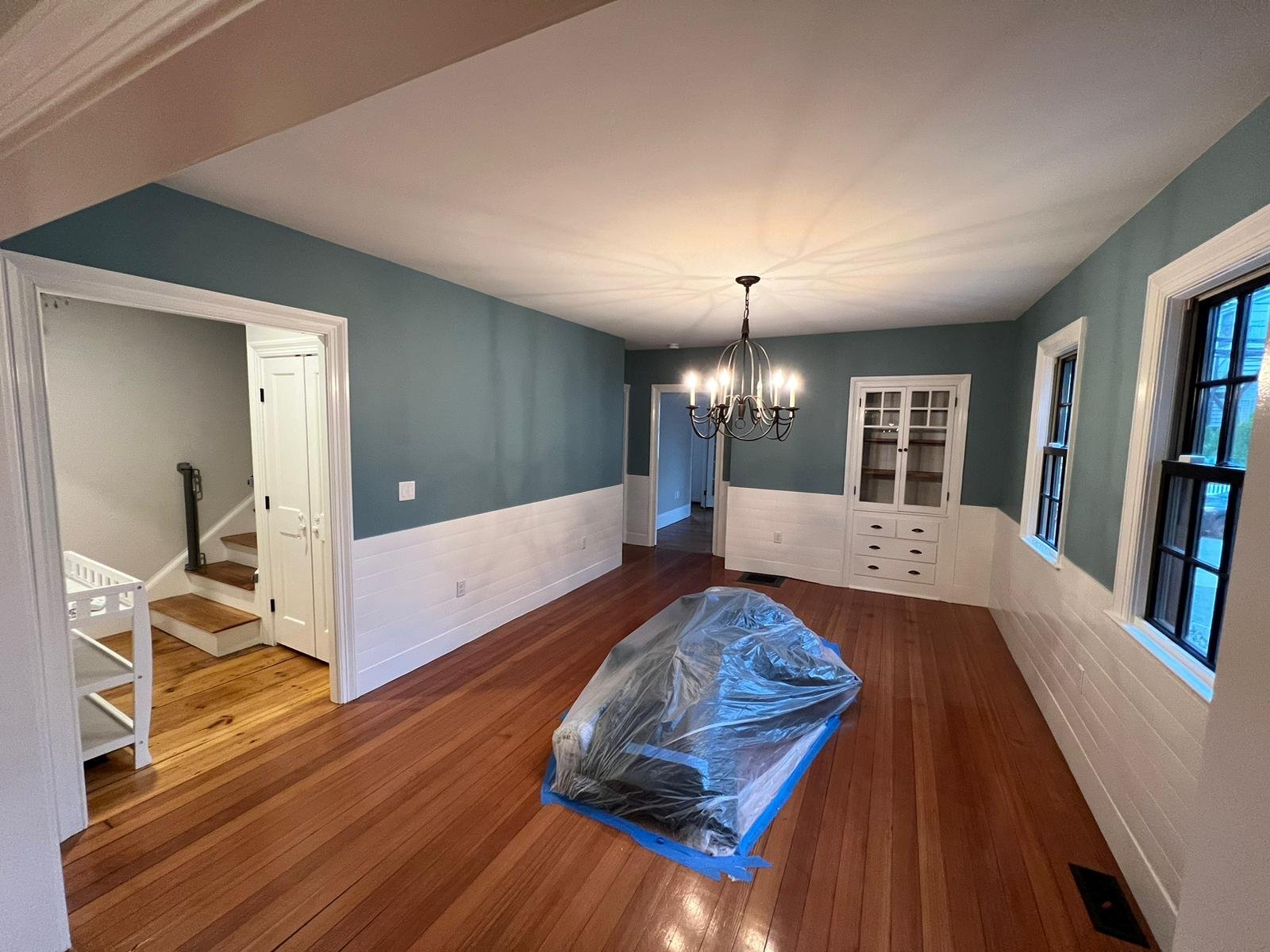 Empty dining room with hardwood floors, blue walls with white wainscoting, large windows, a chandelier, and a covered piece of furniture or appliance on the floor in Chestnut Hill, Boston.