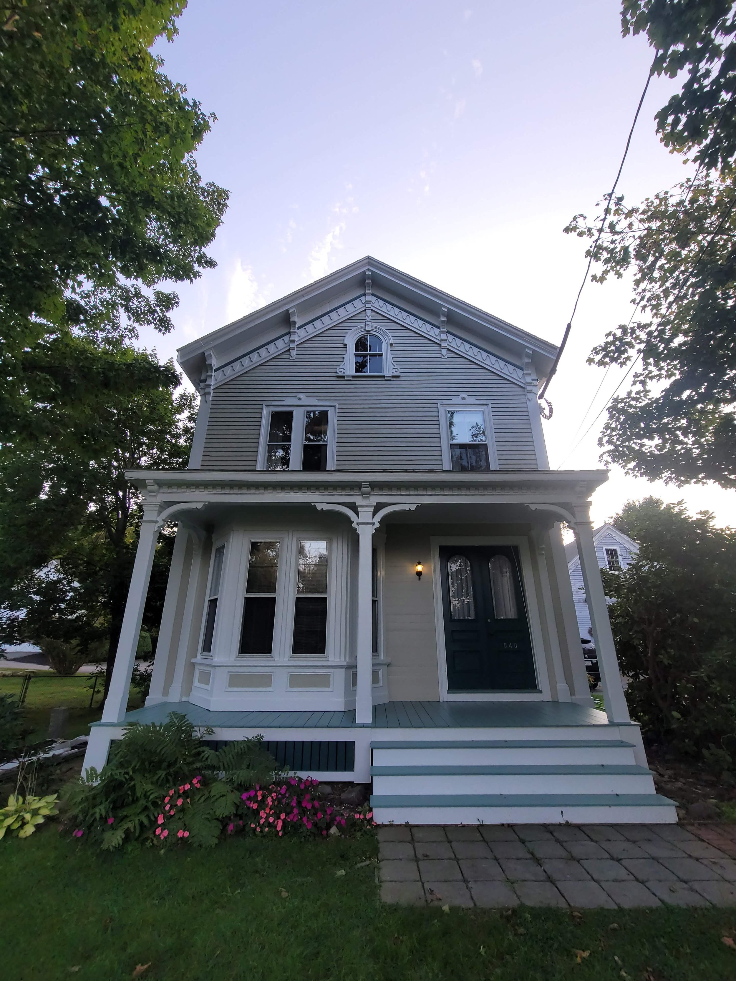 A Victorian-style house in Randolph, Massachusetts with white trim and blue exterior, featuring a front porch with steps, surrounded by grass, flowers, and trees, under a clear sky.