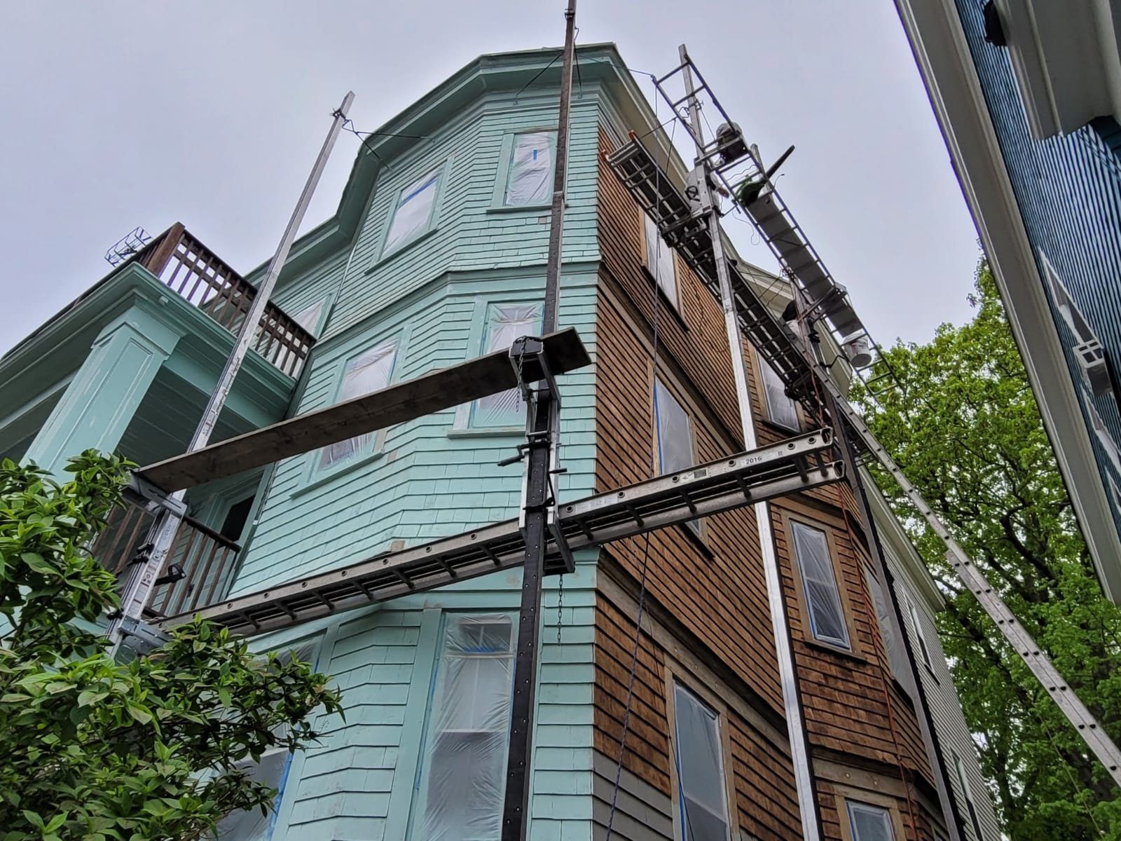 Exterior of a multi-story house under painting project, with scaffolding and construction materials around it. The house has light green primer on one side and refinished wooden siding on the other, with several windows covered in plastic.