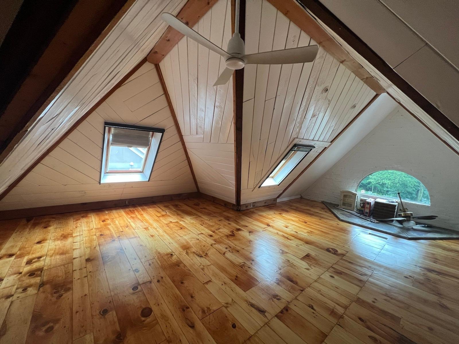 Empty attic room with floor refinishing, angled painted ceiling with two skylights, and a small arched window with a view of trees outside, with a ceiling fan hanging from the ceiling on the Jamaicaway, Boston.