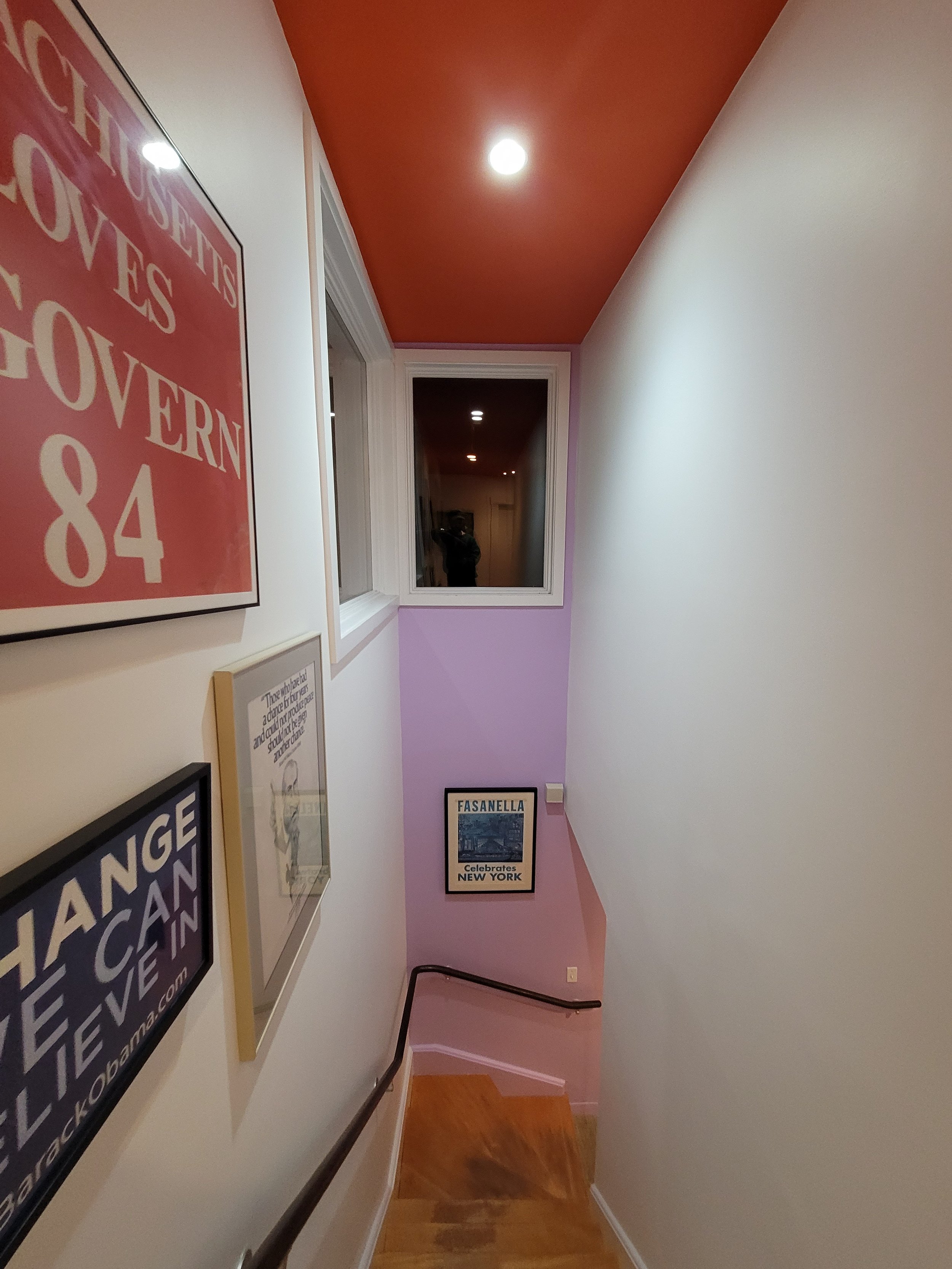 A staircase with framed posters on a wall, including a red poster with white text and a smaller poster with Boston themed images, leading down to a landing with a purple wall and a window in Cambridge, Massachusetts.