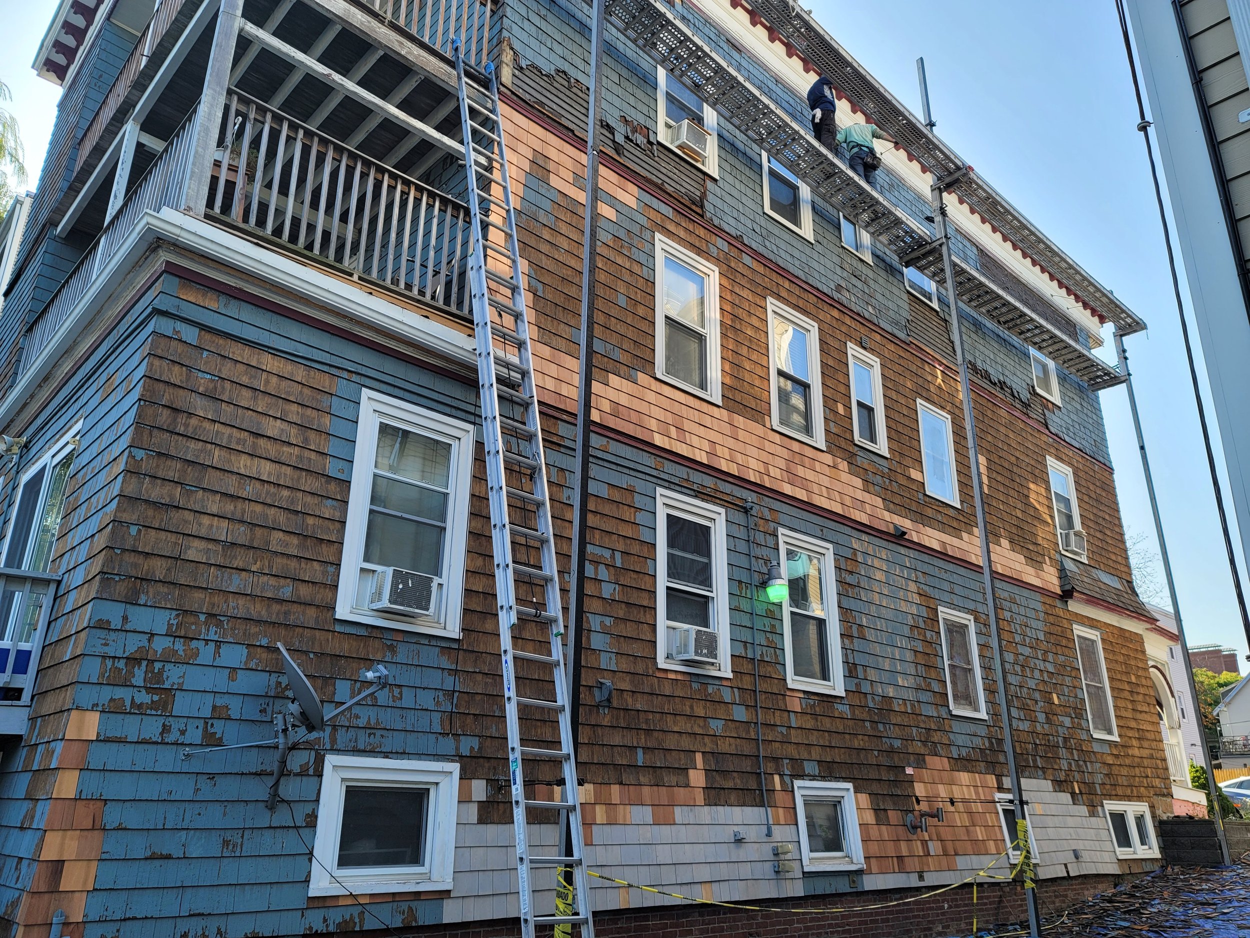 Ryan Painting crew members carrying out a carpentry and restoration, exterior painting project in Dorchester, the has newly installed shingles, peeling blue and brown paint and multiple windows with air conditioning units.