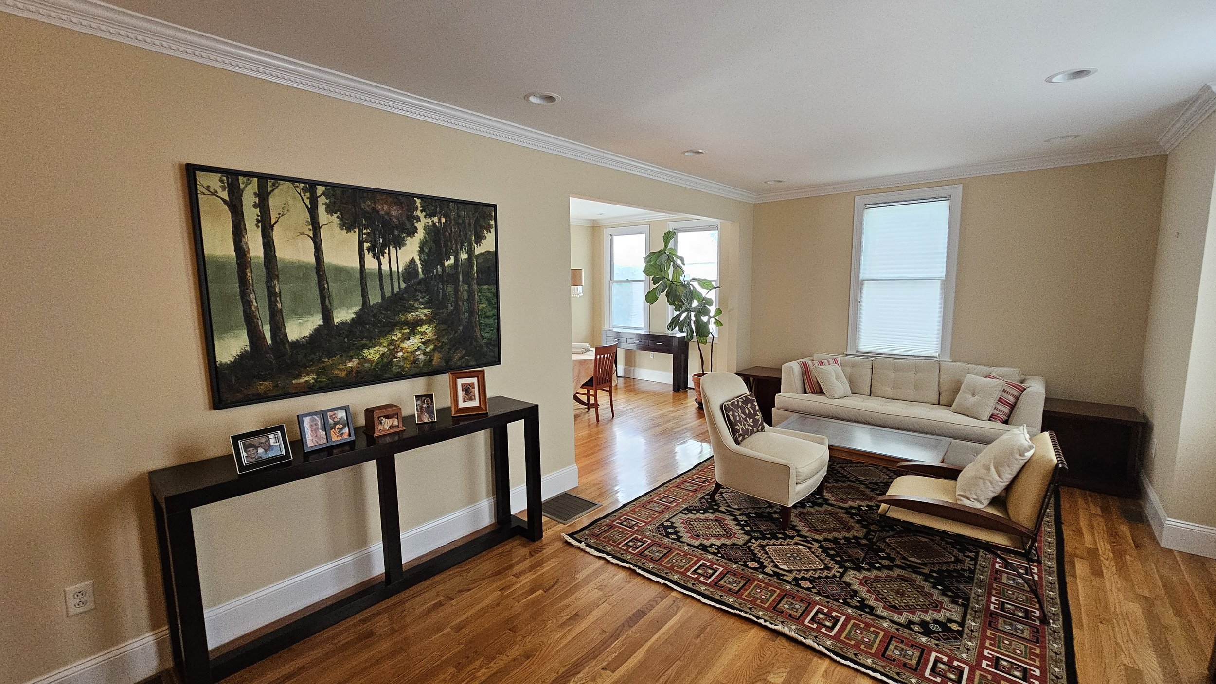 A stunningly repainted Bakersfield Street, Dorchester living room with beige walls, hardwood floors, a large painting of trees by a river, a black console with framed photos.
