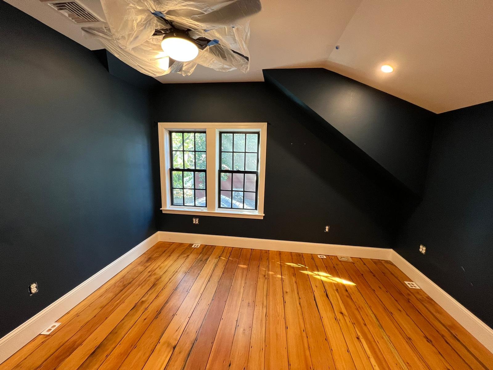 Empty room in a colonial-style Dorchester home with dark blue walls, a window with white trim, hardwood floors, and a ceiling. Part of the ceiling appears under renovation, covered with plastic and tape by Ryan Painting.