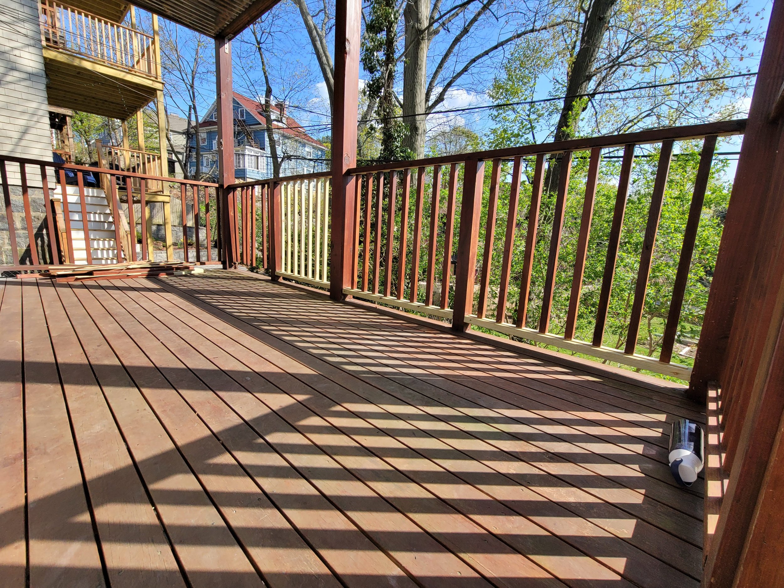 Empty wooden porch with railing, recently deck stained by Ryan Painting, shadows cast by railing, trees and houses in the background.