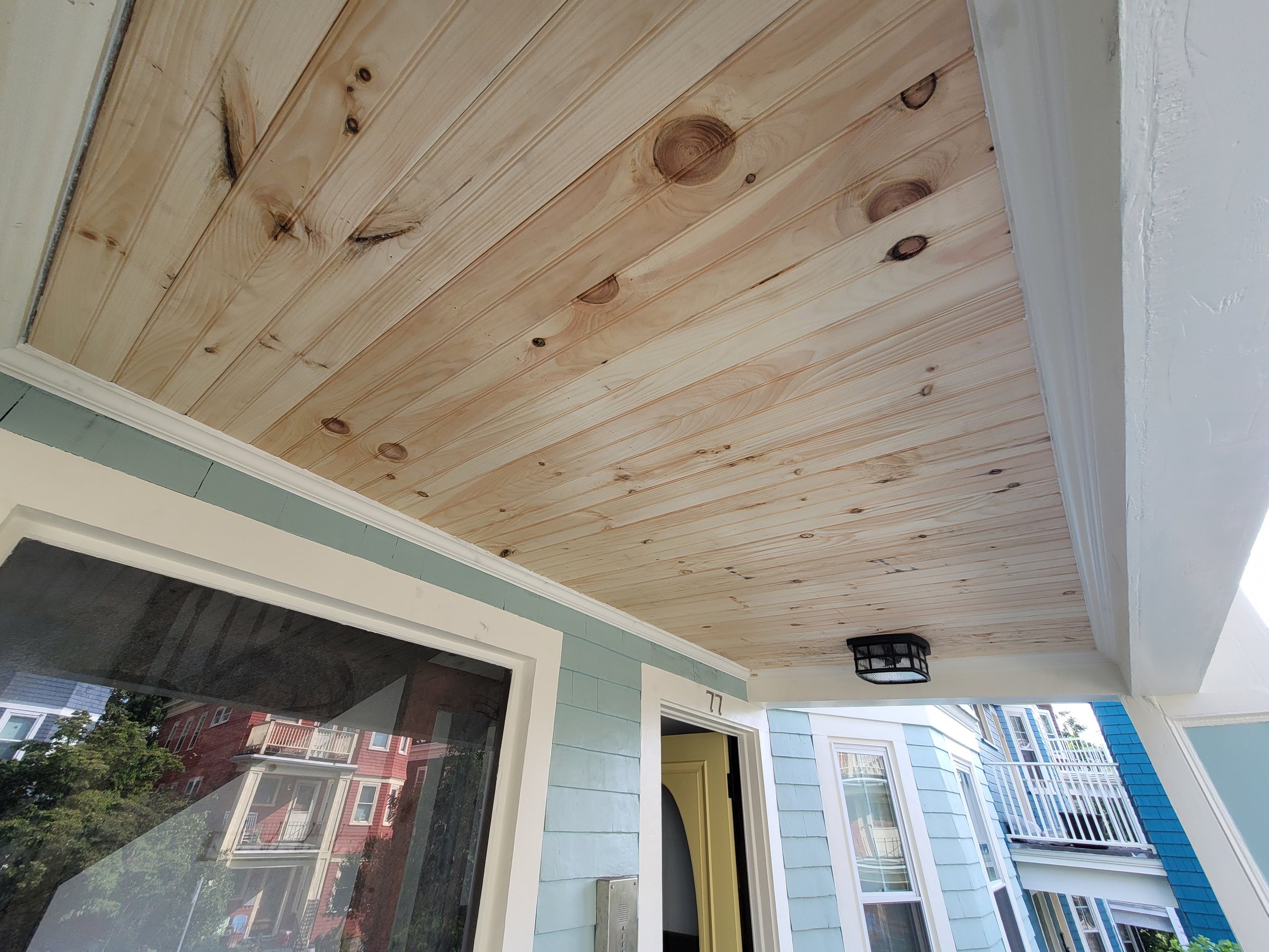 Porch ceiling with newly installed wood planks, light blue house exterior, window, and partial view of neighboring houses on Montebello Street, Jamaica Plain.